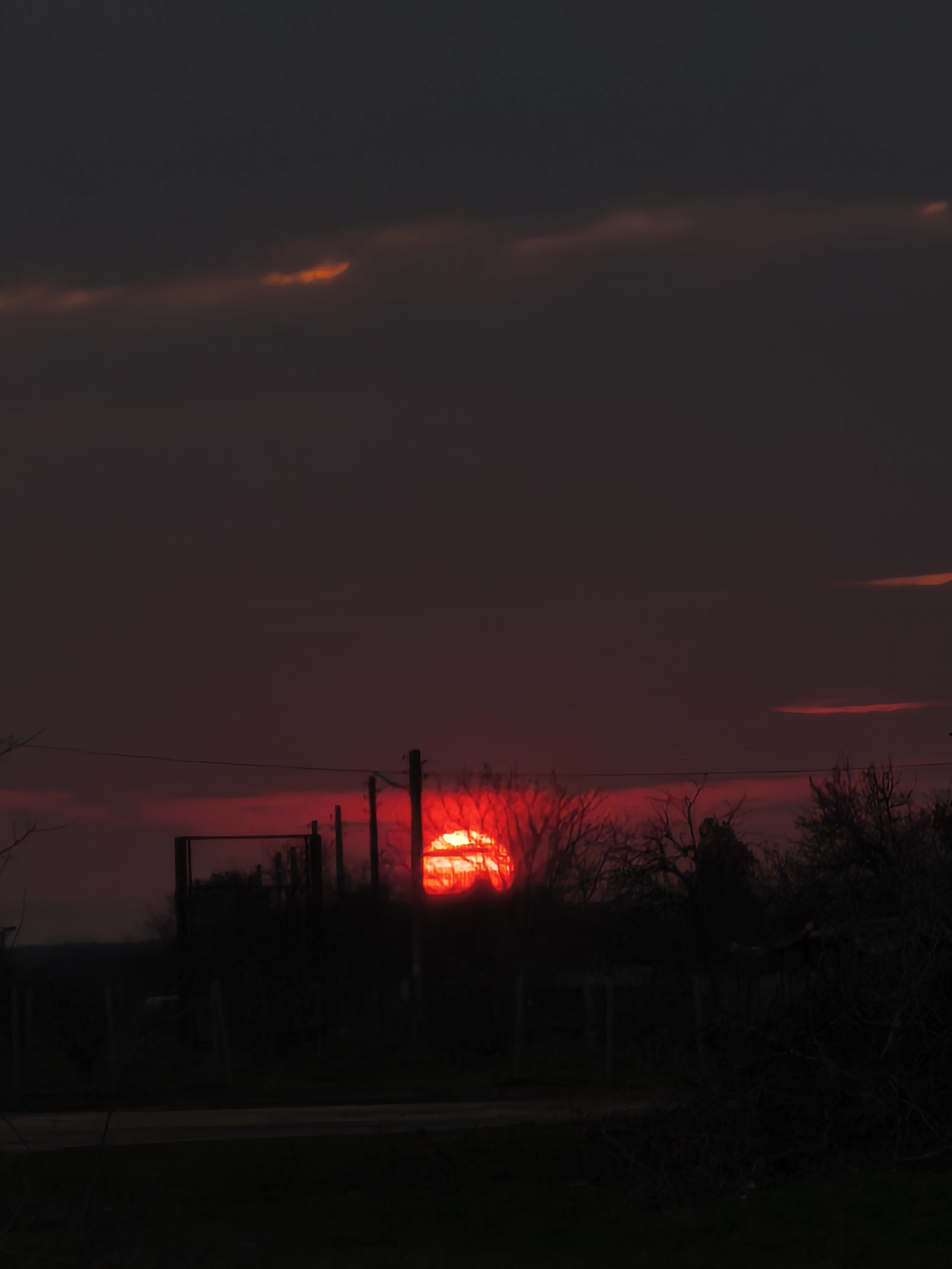 The image captures an outdoor scene in Slavyanovo, Bulgaria, under low light conditions consistent with either dusk or dawn. A prominent, intensely red, circular solar disk is positioned low on the horizon, partially obscured by a dense layer of dark, silhouetted vegetation, primarily consisting of bare trees and shrubs with visible branches. The sky above the horizon is a deep purplish-gray, with several horizontally oriented, elongated clouds exhibiting faint orange and red illumination, most notably in the upper left and upper right quadrants. In the midground, multiple dark utility poles stand vertically, supporting horizontal lines that span across the scene. To the left of a central utility pole, a dark, rectangular, open-framed structure is discernible. The foreground consists of dark, undifferentiated ground, which appears to include a dark, horizontal strip resembling a road or path, along with further silhouetted vegetation and ambiguous fence-like structures. No individuals or discernible actions are present in the scene.