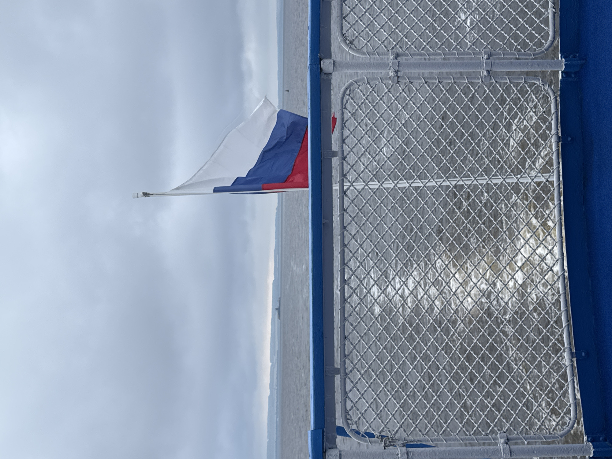 The image captures a view from the deck of a boat on a cloudy day. A Russian flag is prominently displayed on a flagpole, partially obscuring a vast, grey expanse of water and a hazy, distant shoreline. The foreground features a railing with blue and white accents, and a chain-link fence that separates the deck from the water. The water shows a visible wake, indicating the boat's movement. The location is the city of Laishevo, Russia.