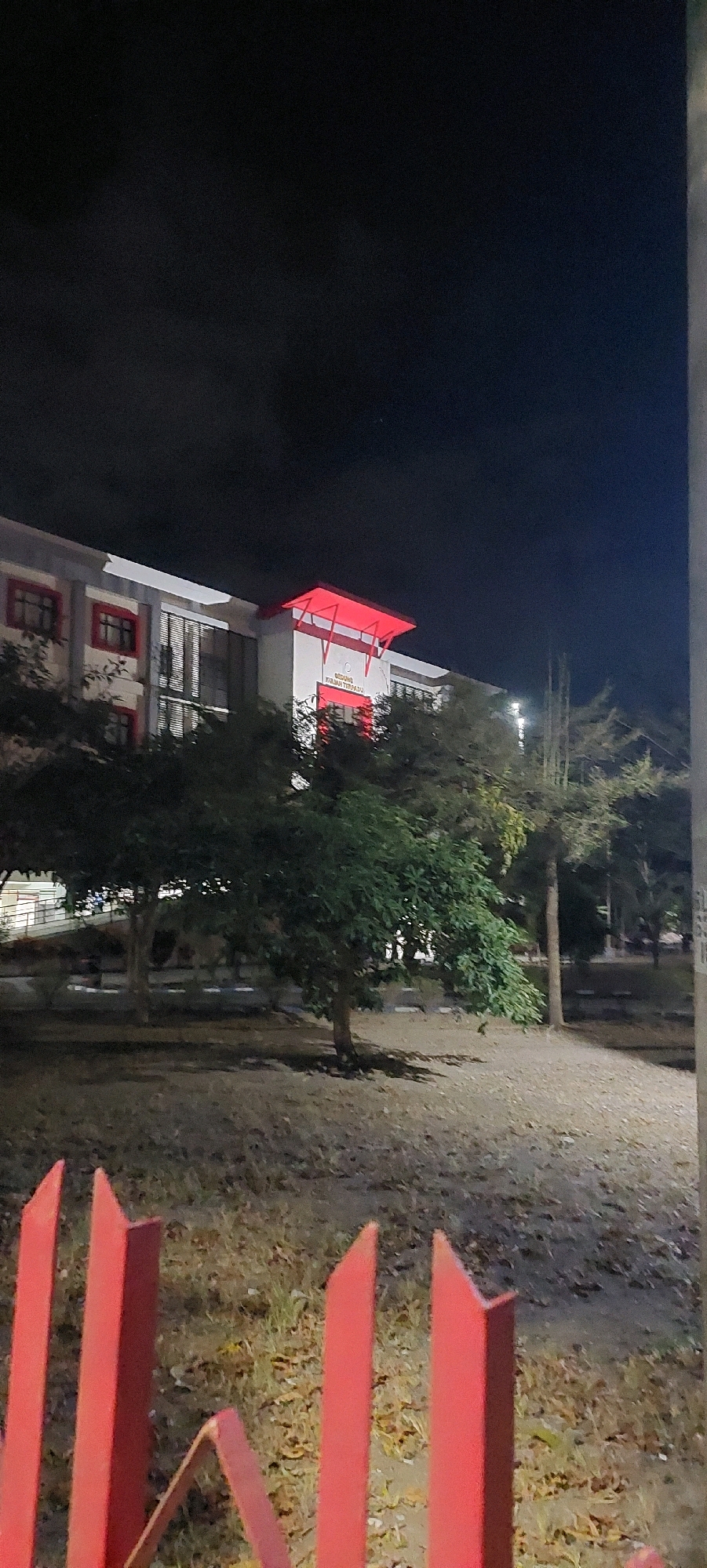 The image depicts a building at night, illuminated by a red canopy and interior lights. The building appears to be a public institution, with signage visible on its facade. In the foreground, there is a red fence, and beyond that, trees and a grassy area with fallen leaves. The overall scene suggests an urban or suburban environment.