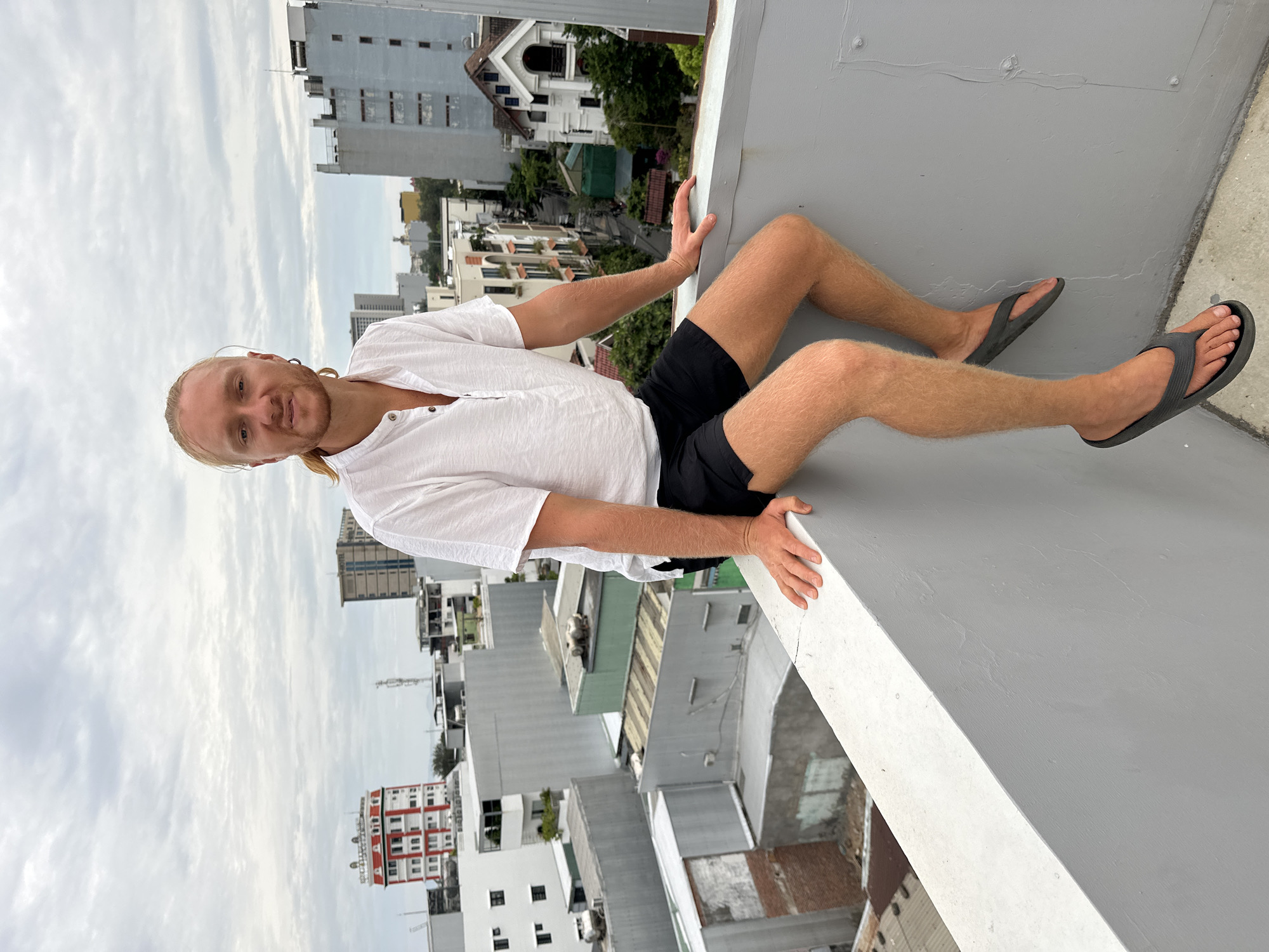 A man with blond hair, wearing a white button-up shirt and black shorts, sits on the edge of a gray concrete rooftop overlooking a city. The man is looking at the camera with a slight smile. In the background, there are several buildings of varying heights and architectural styles, typical of a city in Vietnam. The sky is overcast with clouds. The location is Đà Nẵng, Vietnam.