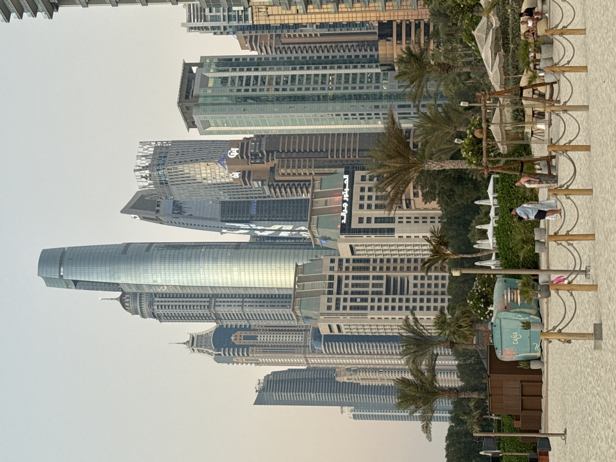 The image shows a panoramic view of the Dubai Marina skyline, a bustling district known for its skyscrapers and luxury developments. In the foreground, a sandy beach is visible, with people relaxing on lounge chairs and enjoying the scenery. A swing set and several umbrellas suggest a recreational area. Palm trees dot the landscape, adding a tropical feel. The background is dominated by a cluster of modern high-rise buildings, their glass facades reflecting the sky. Among them, the distinctive shape of the Princess Tower and the Pier 7 complex are recognizable. The sky is overcast, casting a soft light over the scene, indicating either early morning or late afternoon. The overall impression is one of urban sophistication blended with beachside leisure.