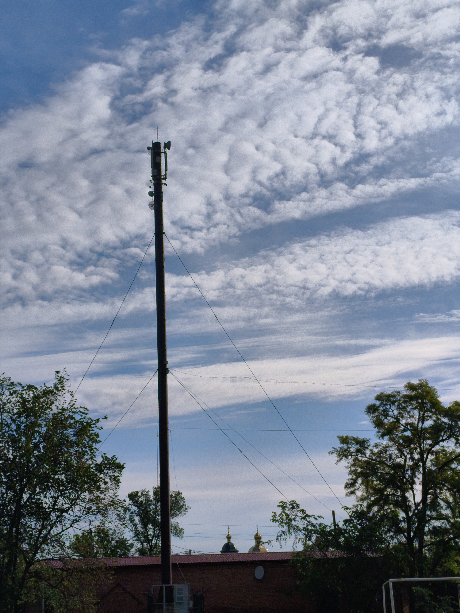 A photograph captures a tall, dark telecommunications pole or mast standing prominently against a bright blue sky filled with white, scattered clouds. The pole, likely made of wood, is equipped with various antennas and dish-like transmitters near its top. Several guy wires extend from the pole, securing it.
In the lower portion of the image, the tops of green trees frame the scene on both the left and right sides. Below the trees and the lower part of the pole, a brick building with a reddish-brown roof is visible. Peeking above the building and trees in the mid-ground are two golden church domes, each topped with a cross, indicative of an Eastern Orthodox church, consistent with the Ustyvytsia, Ukraine location. On the far right, a white structure, possibly part of a goalpost, is partially visible.
The weather appears to be partly cloudy and bright, suggesting it is daytime, likely mid-morning to late afternoon. There is no visible text or discernable human activity. The overall scene depicts a blend of modern infrastructure (the telecommunications pole) and traditional architecture (the church domes) set in a natural, leafy environment.