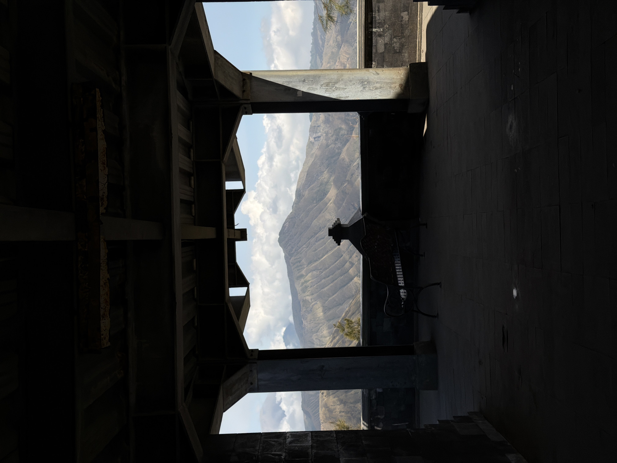 The image captures a view of volcanic mountains from a covered viewing area in Cemorolawang, Indonesia. In the foreground, a dark, open-sided structure with a tiled floor and a dark metal roof creates a silhouetted frame. Within this structure, a dark metal bench with a decorative backrest is visible, facing the panoramic mountain landscape. The mountains themselves are layered and rugged, with visible erosion patterns and some sparse vegetation. The sky is partly cloudy, with bright white cumulus clouds contrasting against a clear blue sky. The lighting suggests it is daytime, with strong shadows cast by the structure onto the floor. The overall scene conveys a sense of stillness and natural beauty viewed from a man-made vantage point.