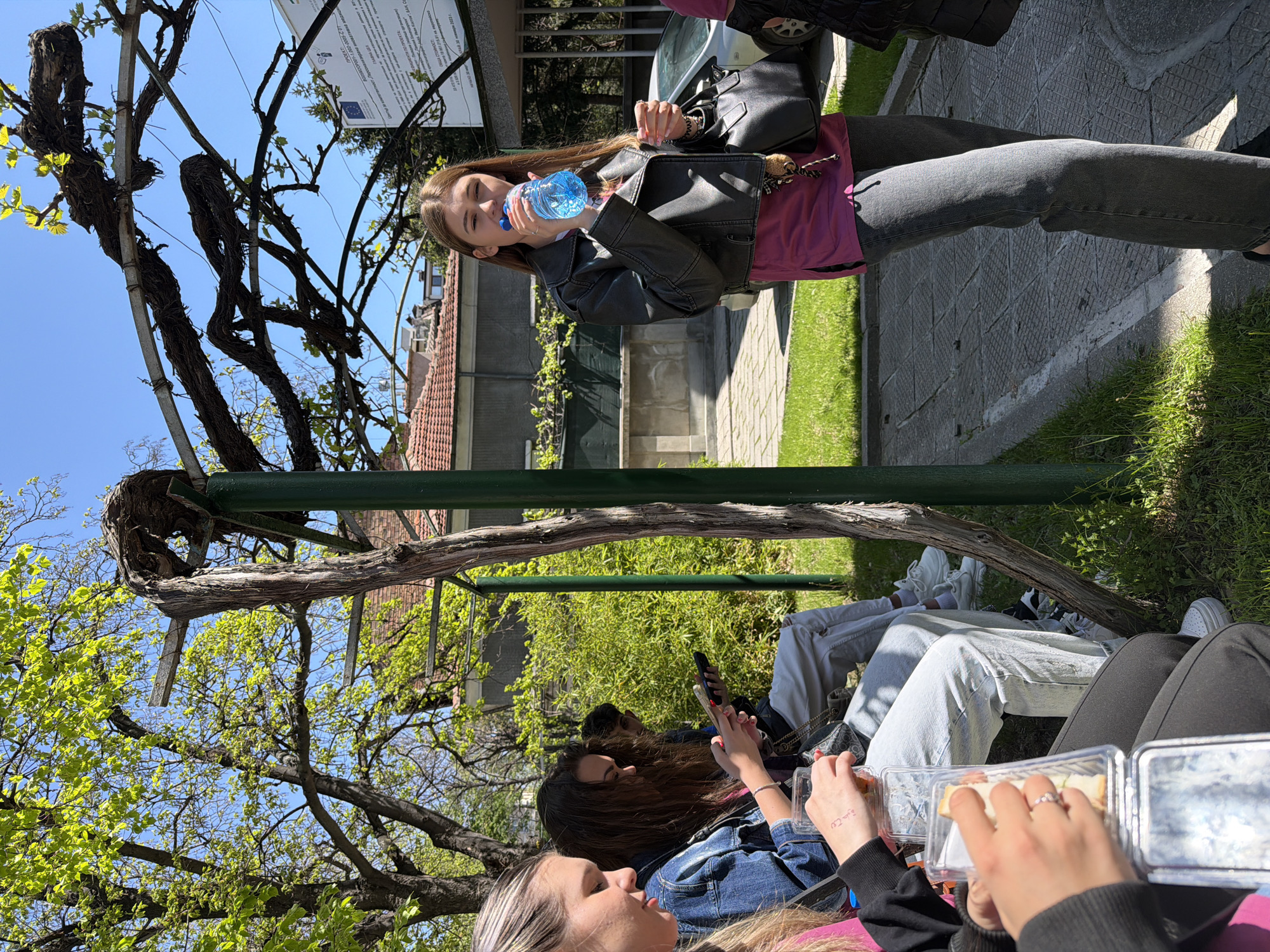 The image captures a casual outdoor scene, potentially a picnic or gathering. At the top of the image, a young woman with long brown hair drinks water from a blue plastic bottle. She wears a black jacket over a pink shirt and grey pants. A black shoulder bag is slung on her shoulder. A car is visible parked behind her.
Below, a group of people is seated, seemingly enjoying a meal or snack. They have food in plastic containers. One person is holding a smartphone. The group appears to be in a park-like setting, with trees and greenery in the background. There's a wooden and metal structure, perhaps a pergola or trellis, providing partial shade.
In the upper left corner, there is a sign with text, including the phrase "Център за компетентност" and details about a project.
Based on the OCR text and the general appearance of the scene, this image might have been taken in or near a park in Plovdiv, Bulgaria, possibly near the city's center of competence.