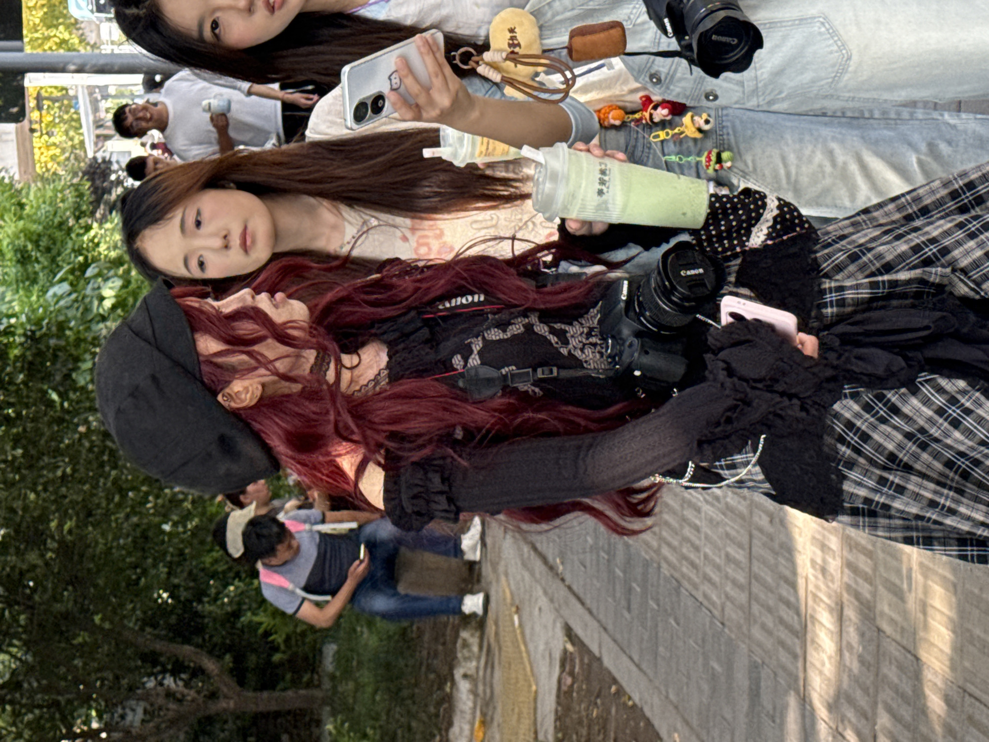 This image depicts a casual outdoor scene in Nan'an District, China, under bright, clear daylight conditions.
The foreground features three young women. One woman with long dark brown hair wears a light-colored top and holds a smartphone in a clear case in her right hand, while her left hand holds a light green beverage in a plastic cup, featuring the visible Chinese text "寻茶记" (Xún chá jì). Another woman with similar dark hair and wearing a denim jacket is partially visible, also looking towards the viewer. The third woman, distinctive with long, wavy dark red hair, wears a black beret, a black intricate top, and a black and white plaid skirt. She carries a black Canon camera with a large lens, displaying the "Canon" brand, and is looking upwards and to her right. The women appear relaxed, engaged in a social interaction, possibly taking photos or enjoying a casual outing.
In the midground and background, several other people are present; one man in a white t-shirt holds a cup and smiles, while another man in a grey and blue t-shirt and blue jeans is seated, engrossed in his phone. The setting includes a paved path or sidewalk and lush green foliage from trees and bushes, suggesting a park or urban pedestrian area. Visible objects include multiple smartphones, drinks, and small keychains or charms, such as a yellow plush item with faint Chinese characters (possibly "今天真好") and cartoon figures. The overall atmosphere is lively and contemporary.