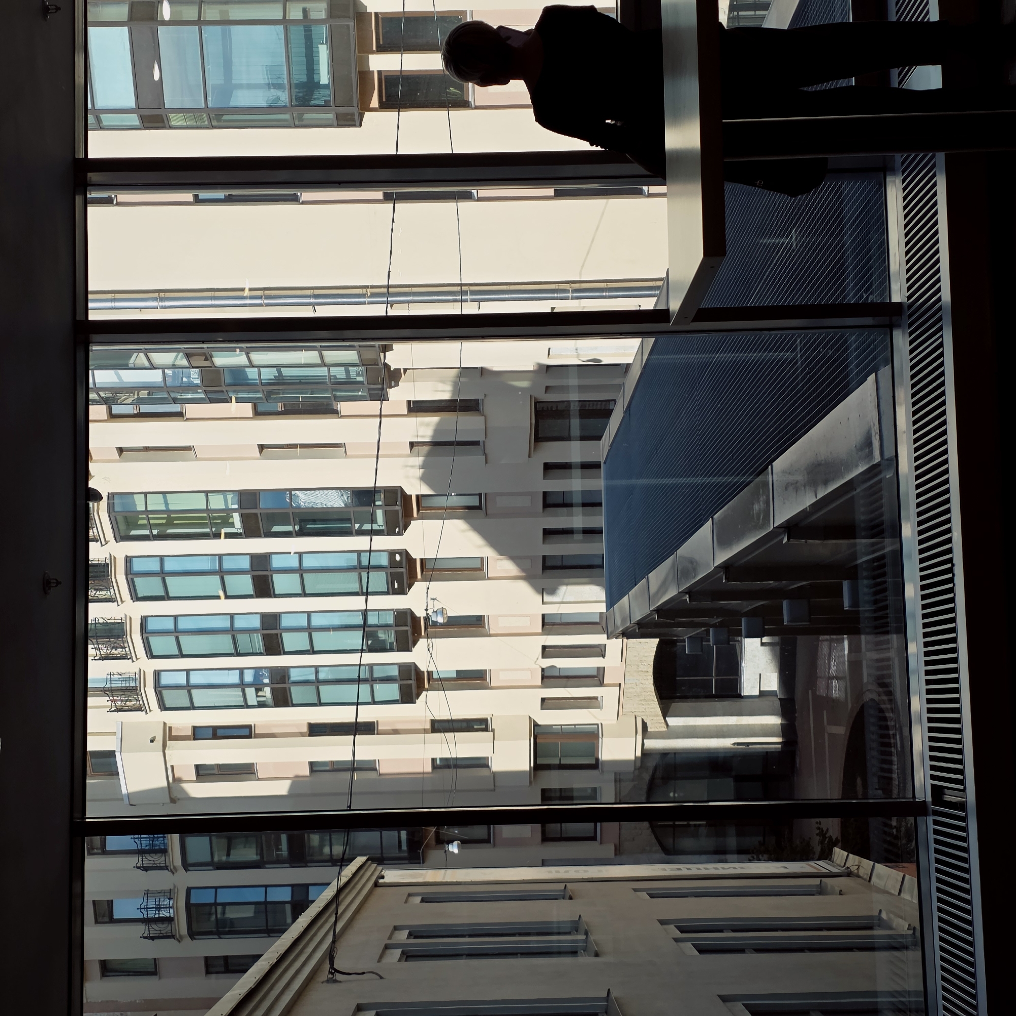 The image captures an urban scene viewed through a large window from an indoor vantage point, likely an upper floor. In the upper right quadrant, a person is silhouetted against the bright exterior, leaning slightly over a window frame or ledge, appearing to look out or down at the buildings below. Their head and upper torso are visible, darkened by backlighting.
The view outside reveals a dense arrangement of city buildings under clear daylight, suggesting either morning or afternoon due to the prominent shadows cast on the building facades. The central building, directly across from the viewer, is multi-storied with numerous rectangular windows that reflect the sky and surrounding structures, giving them a blue-green tint. Its facade is a light beige or cream color with darker accents. To the right, another building features a distinctive modern design with a dark, possibly slatted or textured, roof structure visible through the window. Further down and to the left, another building facade with more traditional windows and balconies is visible. Wires or cables can be seen running down some of the building walls, and a small, light-colored fixture is attached to one facade.
Reflections are a significant element, with architectural details and skies mirroring in the extensive glass surfaces, creating layers of visual information. In the lower right window reflection, faint, possibly Cyrillic text is visible. The scene portrays a bustling urban environment, characteristic of a large city like Saint Petersburg, Russia, emphasizing architectural density and modern design elements alongside more classic building styles.
