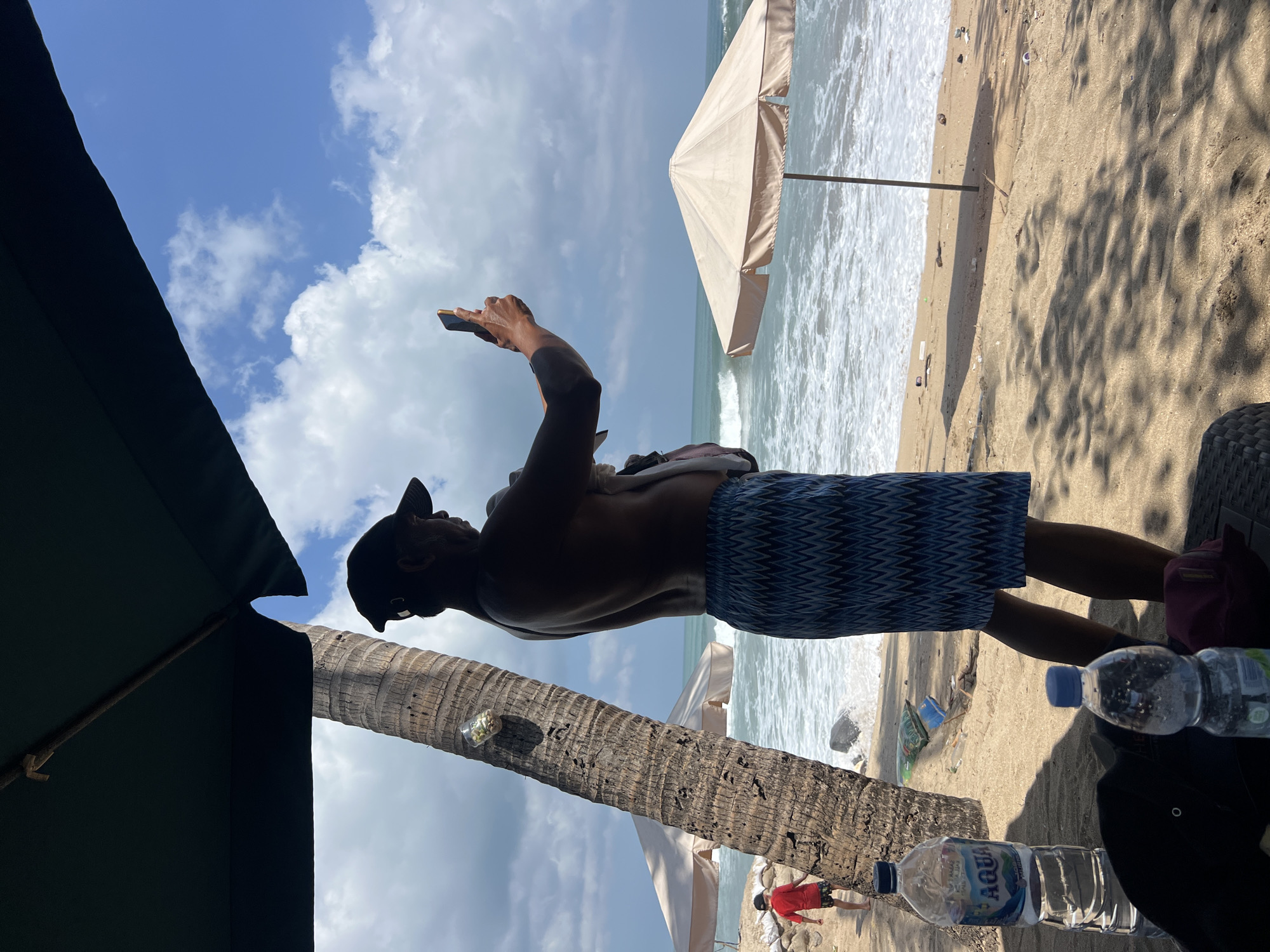 A man in swim trunks and a baseball cap stands on a sandy beach in Kuta, Indonesia, holding a smartphone and appearing to take a photo or video. He is partially silhouetted against the bright sky and ocean. Beside him, a coconut palm tree leans over the scene. In the background, waves crash onto the shore, and beach umbrellas dot the sand. A young child in a red shirt is visible further down the beach. Bottles of water are placed on a surface in the foreground. The scene suggests a typical tourist day at the beach in Kuta.