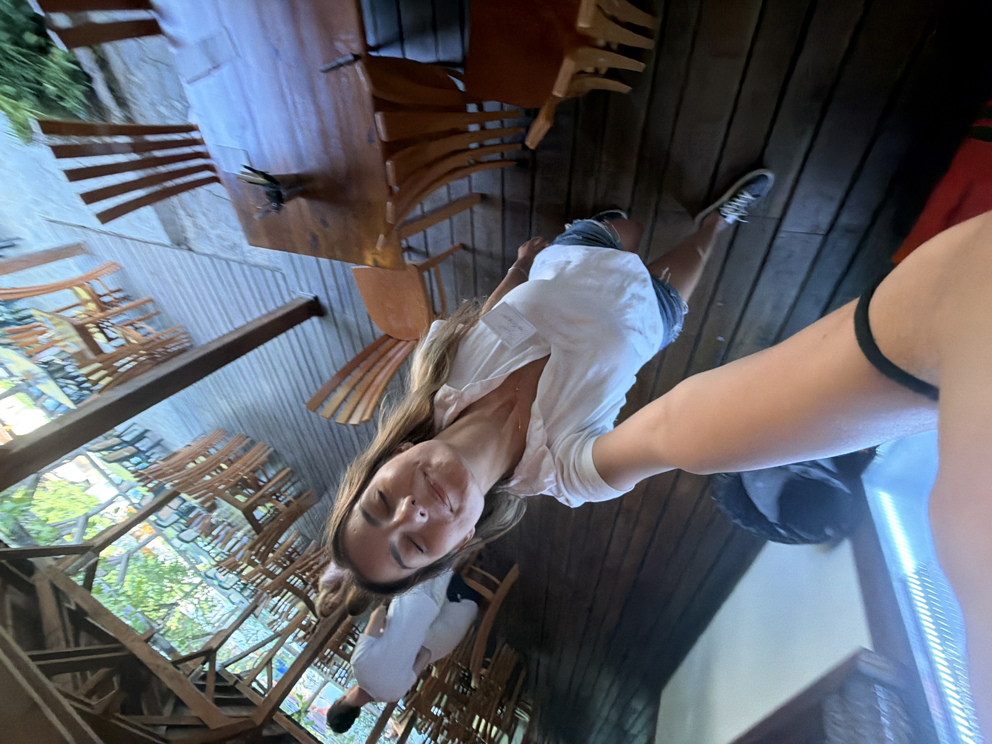 The image shows a woman taking a selfie with her eyes closed and a smile. She is wearing a white t-shirt and denim shorts. In the background, there are wooden tables and chairs, suggesting an outdoor seating area of a restaurant or cafe. A man in a white t-shirt is visible to the left. The environment appears to be a rustic wooden structure with natural foliage visible in the background. The perspective indicates the photograph was taken from a low angle, likely by the woman holding the camera. The scene is set at a restaurant in Tsarevo, Bulgaria, indicated by the presence of a name tag that reads "Restaurant Tsarevo" on the woman's shirt.