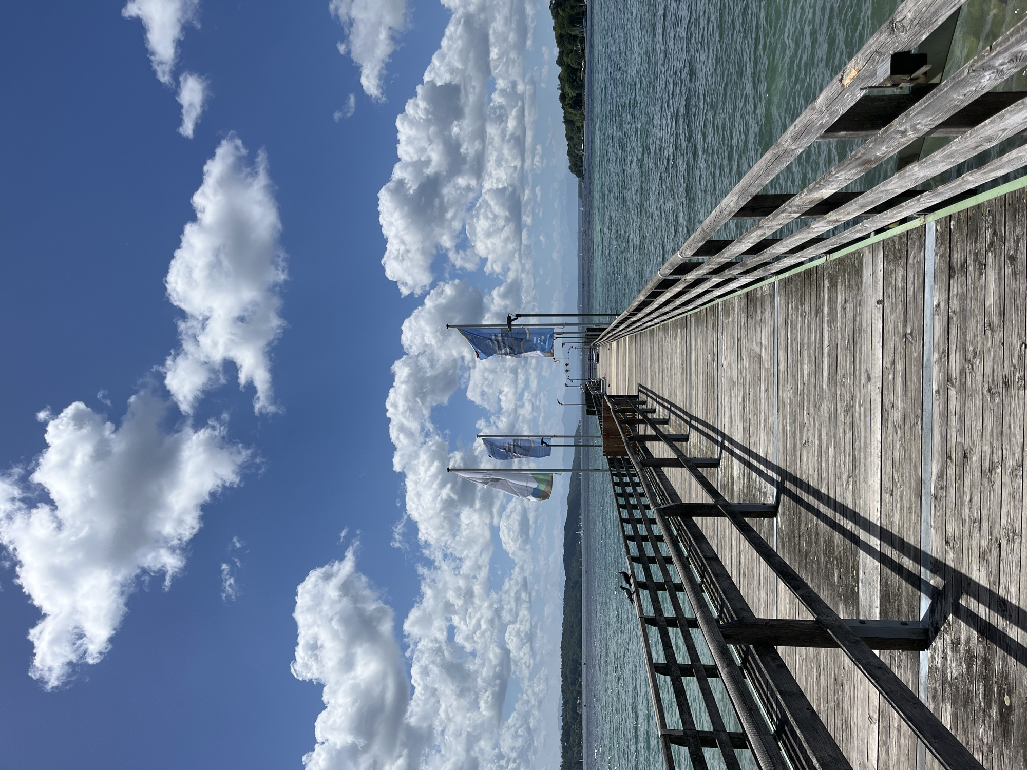 This image depicts a wooden pier extending into a body of water. The pier has a wooden railing on both sides, supported by vertical posts. Two birds are perched on the railing in the foreground. Several flags are planted on the pier in the middle ground, one of which is blue with white text and symbols. The water is a deep teal color and appears to be a lake. In the distance, there is a tree-lined shore and mountains are visible on the horizon. The sky is blue with scattered white cumulus clouds. The scene is likely the Starnberg Lake pier in Starnberg, Germany, a popular tourist destination known for its picturesque views and recreational activities.