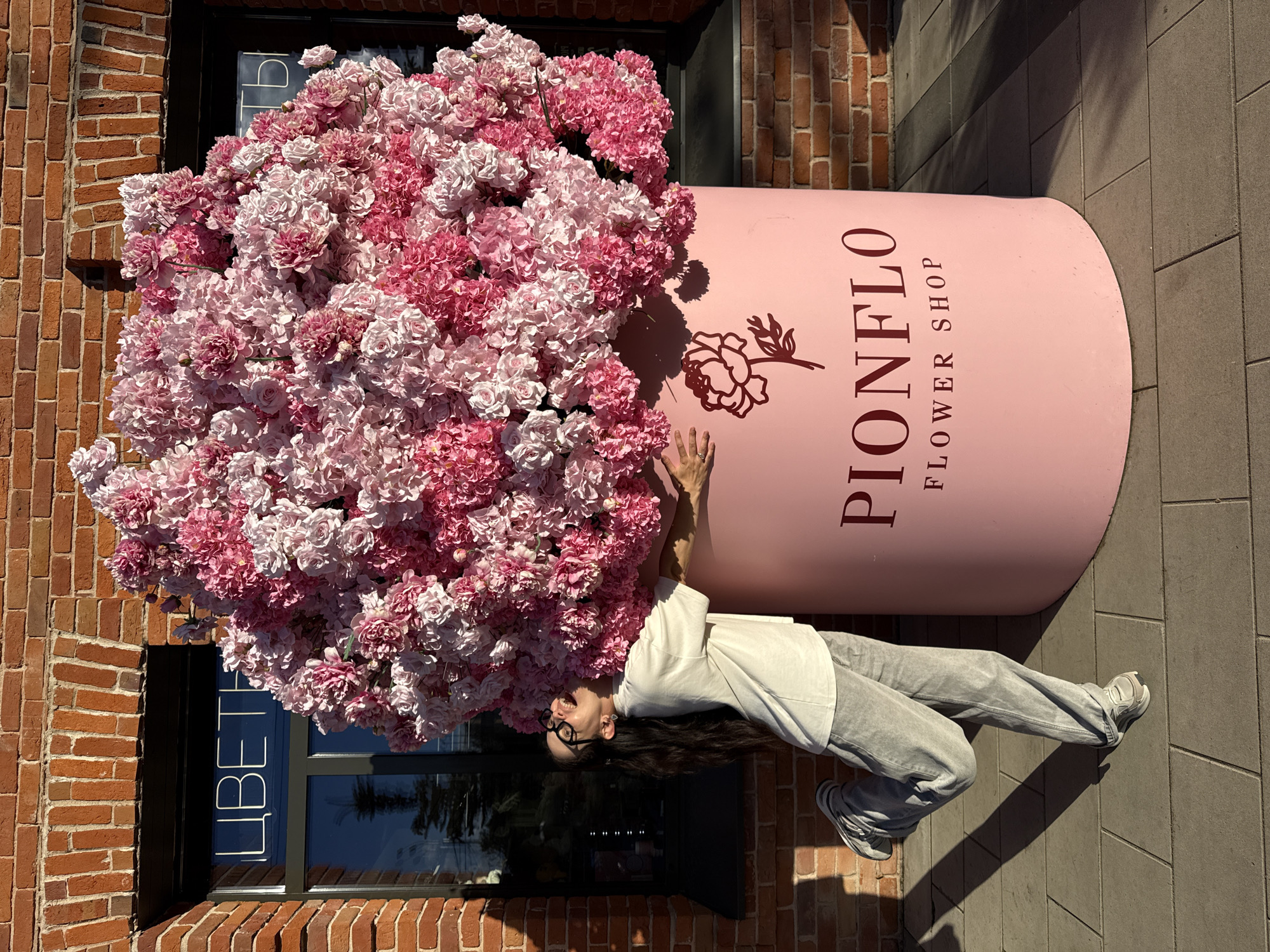 In Kazan, Russia, outside a brick building with arched windows displaying "ЦВЕТЫ" (Flowers) in white neon lettering, a young woman with long dark hair and glasses leans back in playful ecstasy, hugging a colossal arrangement of pink and white artificial flowers. The flowers spill from a large, pale pink cylindrical planter bearing the logo and name "PIONFLO FLOWER SHOP" in dark red lettering. The woman is dressed in a white t-shirt and light grey sweatpants, with grey sneakers on her feet, one leg bent and raised as if in a dance. She is positioned on a paved walkway bathed in sunlight, casting long shadows from the planter and the building.