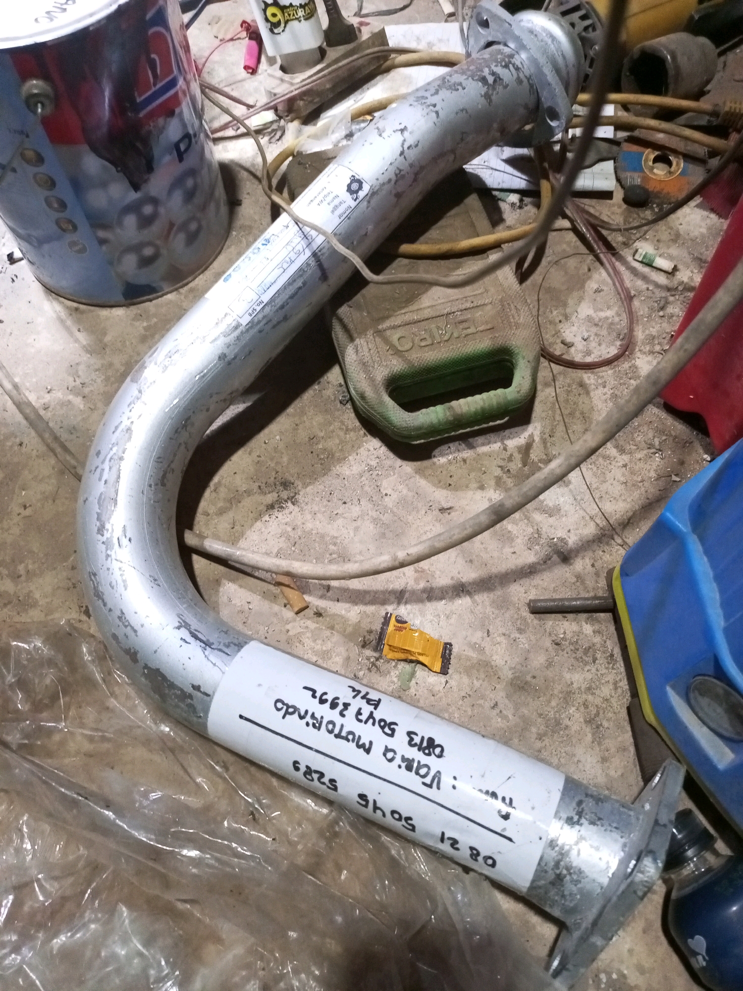 The image displays a silver exhaust pipe lying on a concrete floor, with various objects scattered around it. The exhaust pipe is curved and has a white sticker with black writing on it, which appears to be a product label. Nearby, there is a partially visible blue plastic container, a green jerrycan-like object with a handle, and a red object that could be another container or piece of equipment. Several electrical wires and cables are also present, along with a metal can with a red and blue label, and a small yellow packet. The overall environment suggests a workshop or storage area, potentially in Keling Panggau, Indonesia, given the context. There are no people visible in the image, and no actions are being performed by any subjects.
