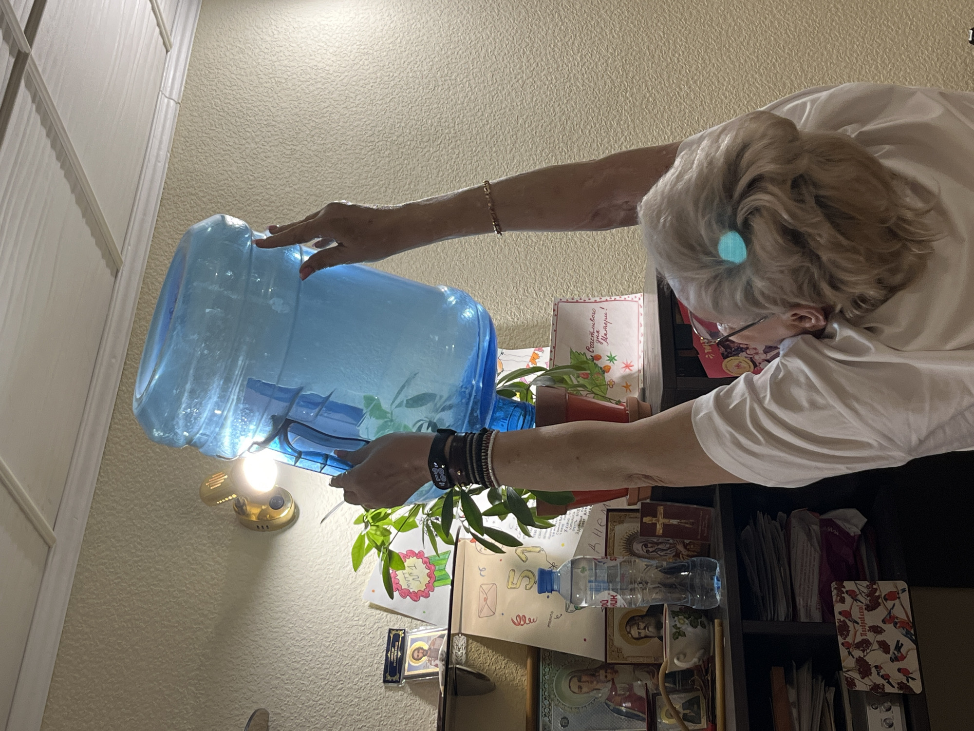 An elderly woman with short blonde hair is pouring water from a large blue water bottle into a potted plant. She is wearing glasses and a white t-shirt. The plant is sitting on a shelf among various items including religious icons, books, greeting cards, and another bottle of water. A light fixture is visible on the wall behind the water bottle. The specific location of this scene within the city of Chaikovsky, Russia, is not discernable from the image.