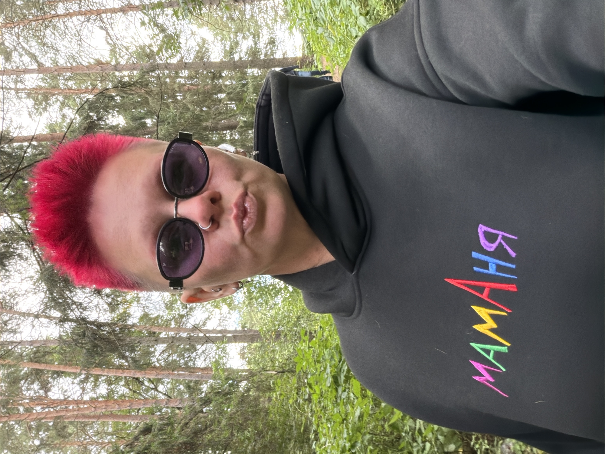 A person with bright pink, spiky hair and oval sunglasses poses for a selfie outdoors. They are wearing a black hooded sweatshirt with the word "МАМАня" embroidered in colorful letters. The background consists of a forest with tall trees and green foliage. The person is pouting their lips towards the camera and wearing hoop earrings. The scene is likely located in Chaikovsky, Russia, given the context provided. There are no discernable interactions with others, and the focus is on the individual.