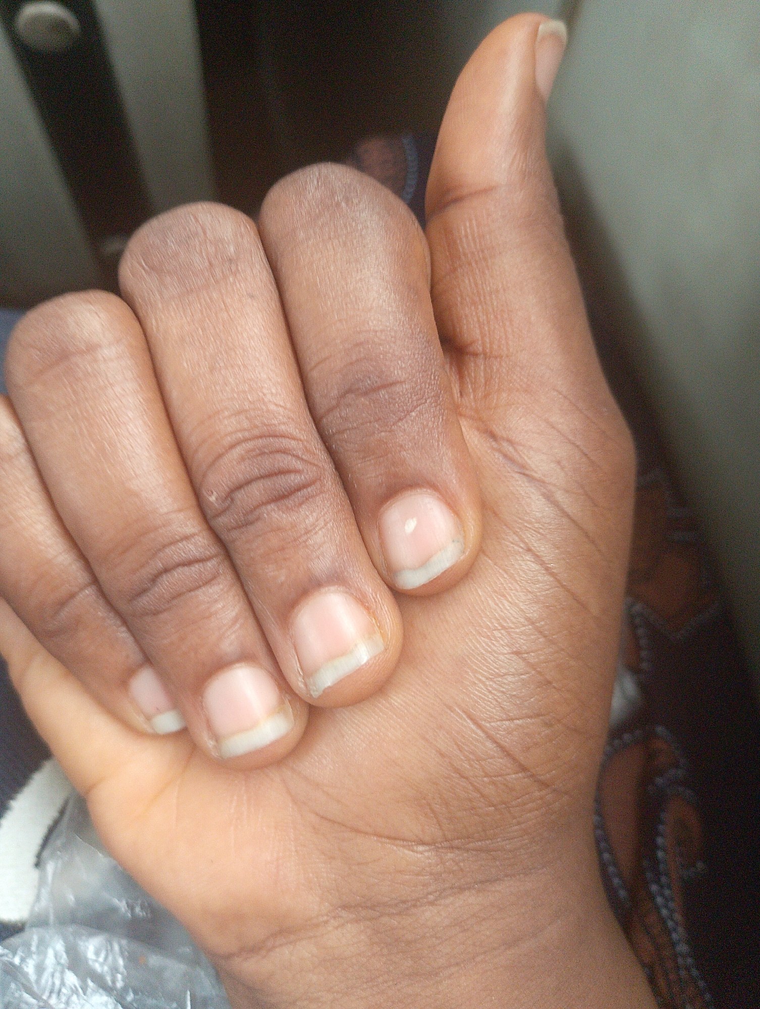 A close-up, top-down view shows a dark-skinned person's left hand, positioned on a patterned surface that appears to be fabric. The fingers are slightly curled inward, with the thumb extended upwards. The fingernails are short and appear clean, with a faint white crescent visible on some of them. The skin on the hand displays natural lines and creases. In the background, out of focus, is a vertical object with a circular element at the top, possibly a light fixture or part of a door frame, and a textured, muted green surface. The overall lighting is dim, with a slight glare on the fingernails. The scene is indoors, in a location that has not been identified as being in Sabojibi, Nigeria.