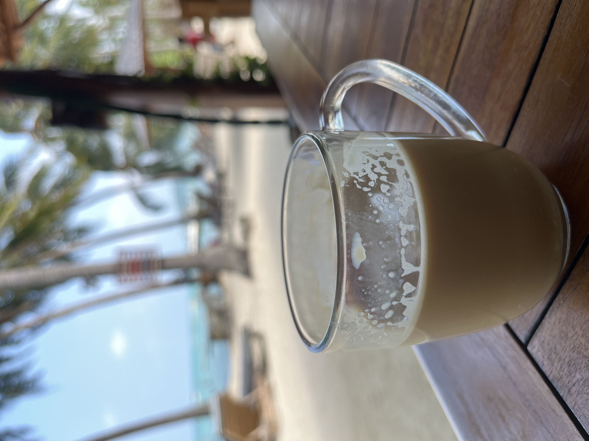 A close-up, eye-level shot shows a glass mug of light brown liquid, likely coffee with milk, resting on a wooden table. The mug has a clear glass handle and shows remnants of foam clinging to the inside rim and walls. The table's surface is made of parallel wooden planks. In the background, a blurred beach scene unfolds under a clear blue sky with scattered clouds. Palm trees with visible fronds frame the view, and lounge chairs are visible on the sandy beach. The ocean is a turquoise blue. The overall setting suggests a tropical or resort location.
