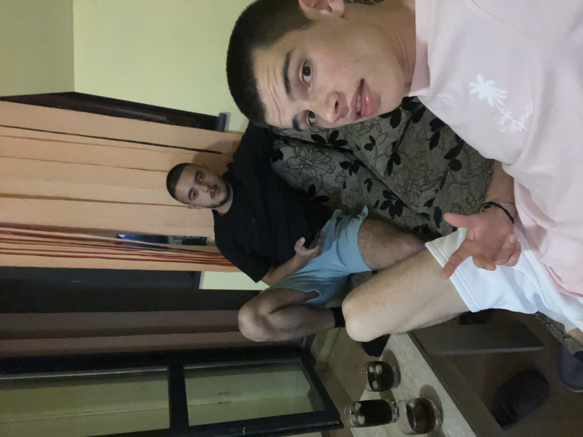 Two men are present in an interior space. One man is seated on a couch with floral upholstery, wearing a pink shirt with a "Santa Monica" logo. The other man is seated next to the couch, wearing a black t-shirt and light blue shorts, holding a phone. Three glasses with dark liquid and ice cubes are on a surface near the men, and a shoe is next to the glasses. A window is partially visible. The scene is likely in a home setting, possibly in Plovdiv, Bulgaria, but without landmarks or specific features, the location is indeterminate.