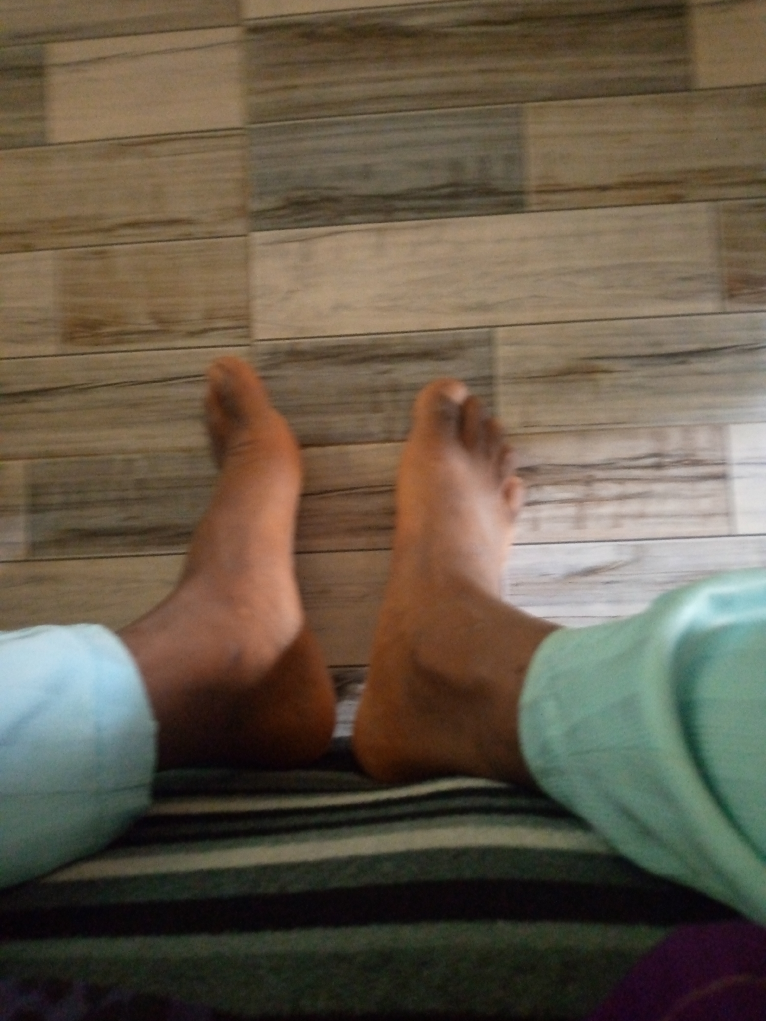The image shows a close-up view of a person's bare feet and lower legs against a backdrop of wooden floor planks. The person is wearing light blue pants. The lighting suggests an indoor setting. The image does not provide enough information to determine the specific location in Jalingo, Nigeria.