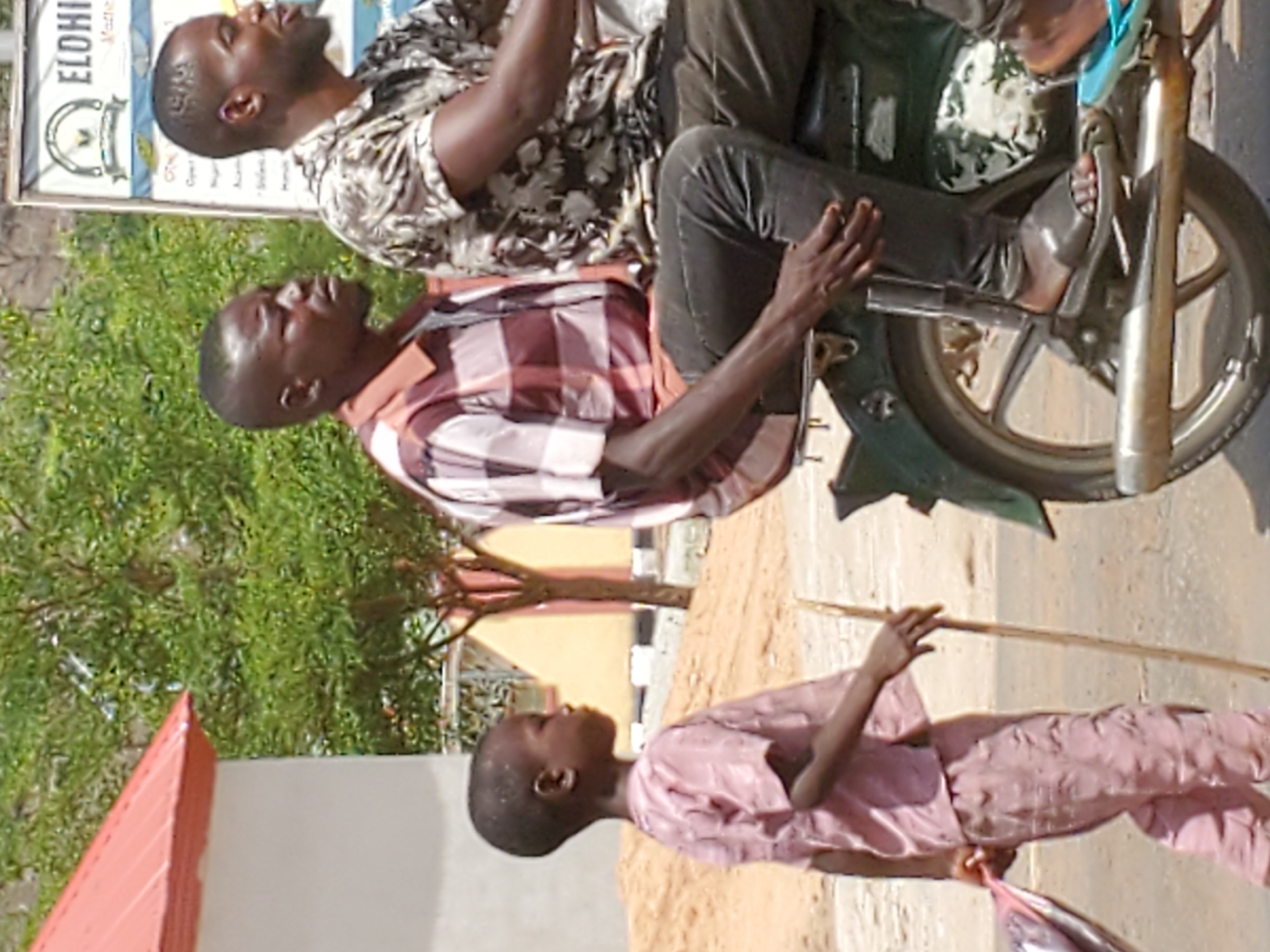 The image shows two men riding a motorcycle on a street in Mararaba, Nigeria. The first man, wearing a black and white patterned shirt, sits behind the driver, who wears a plaid shirt. The motorcycle has a green frame and a silver exhaust pipe. A child in pink clothing stands on the side of the road holding a long stick. A building with a red roof is visible in the background. There is a sign in the background with the word “ELOHI”. A tree can be seen behind the sign. The road appears to be unpaved.