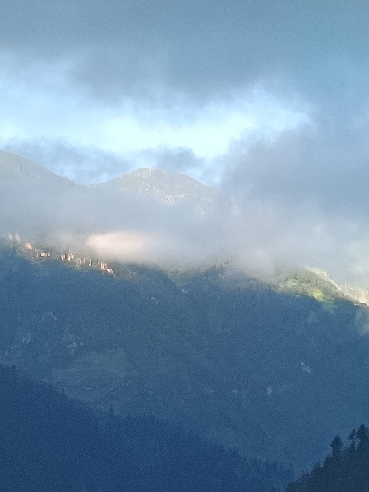 A mountain range is shrouded in mist and clouds. The peaks are dark and covered with trees, with some areas illuminated by sunlight breaking through the clouds. There are patches of green visible on the mountainsides, suggesting vegetation. The sky above is a pale blue, also partially obscured by clouds. The image appears to be a landscape photograph taken in a mountainous region, possibly in or near Vashisht, India, known for its natural beauty and spiritual sites.