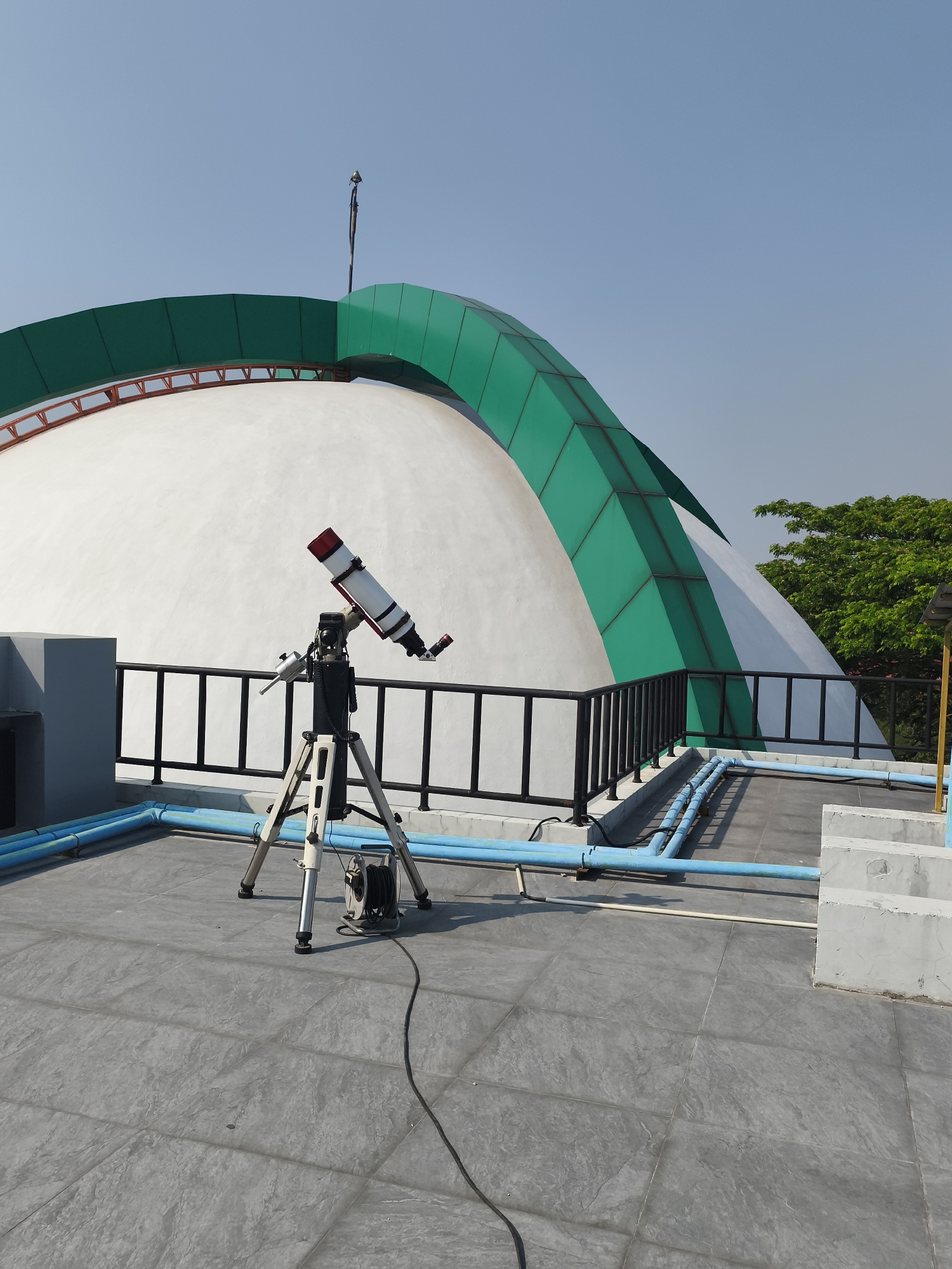 The image shows a white dome-shaped building with green accents, possibly an observatory or planetarium, under a clear blue sky. In the foreground, a telescope on a tripod is set up on a grey tiled rooftop. Blue pipes run along the rooftop. The scene is set in Semarang, Indonesia, as evidenced by the architecture and vegetation. There are no people visible in the image.