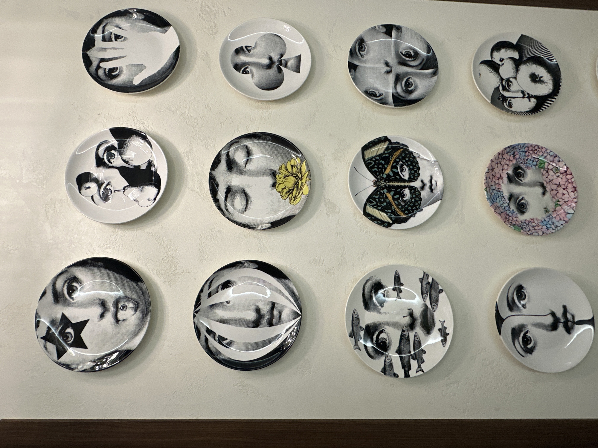 The image displays a collection of decorative plates, each featuring a unique artistic design, predominantly abstract or surrealist portraits. The plates are mounted on a textured off-white wall. The arrangement is in a grid formation, with three rows and three columns of plates visible, plus an additional plate partially visible at the bottom left.
The top row features:
- A plate with a black star over one eye and a piercing on the tongue of a face.
- A plate with two faces, one partially obscured by a pear-like object, and several eyes.
- A plate with a face partially covered by a hand.
The middle row features:
- A plate with a face that appears to be segmented or distorted by vertical lines.
- A plate with a face with closed eyes and a yellow flower in its mouth.
- A plate with a face incorporated into the design of a spade playing card symbol.
The bottom row features:
- A plate with a face where several small fish are swimming around its eyes and mouth.
- A plate with a face wearing a butterfly mask.
- A plate with multiple eyes and faces superimposed on each other.
Below this grid, two more plates are visible:
- A plate on the bottom left with a face that has a dark, pointed shape emerging from its chin.
- A plate to its right with a face surrounded by pink and blue hydrangea flowers.
- A plate to the far right of that with multiple faces and circular shapes.
The lighting suggests an indoor setting, possibly a gallery, store, or home. There are no people or actions depicted, only the static display of the plates. The overall aesthetic is avant-garde and artistic.