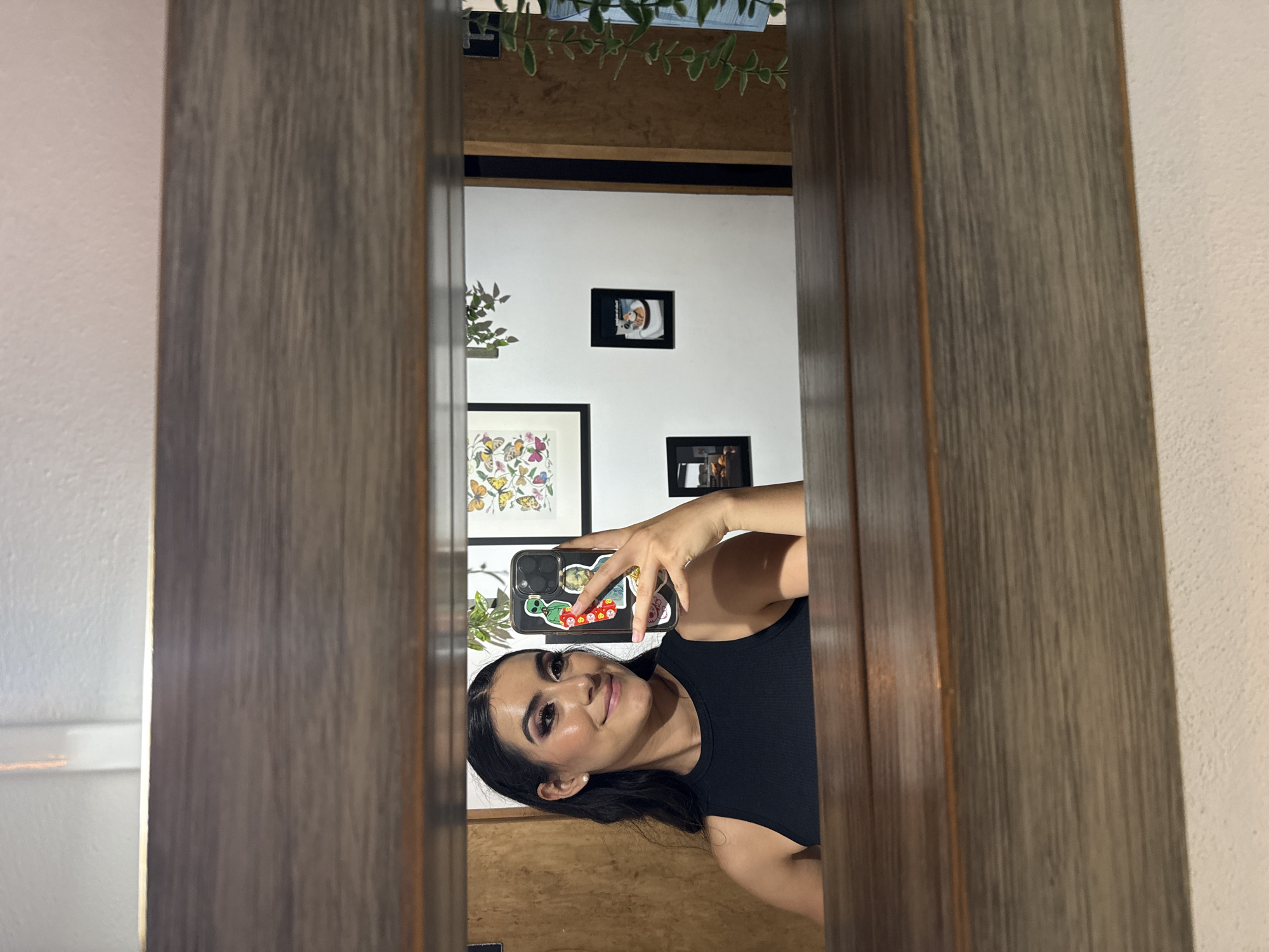 A young woman is taking a selfie in a mirror. The mirror has a dark wood frame. The woman has dark hair and is wearing a black top. She is holding a phone with stickers on the back. The room reflected in the mirror has white walls and several framed pictures. A plant is visible above the frame. The exact location in Puebla, Mexico, is not identifiable from the image.