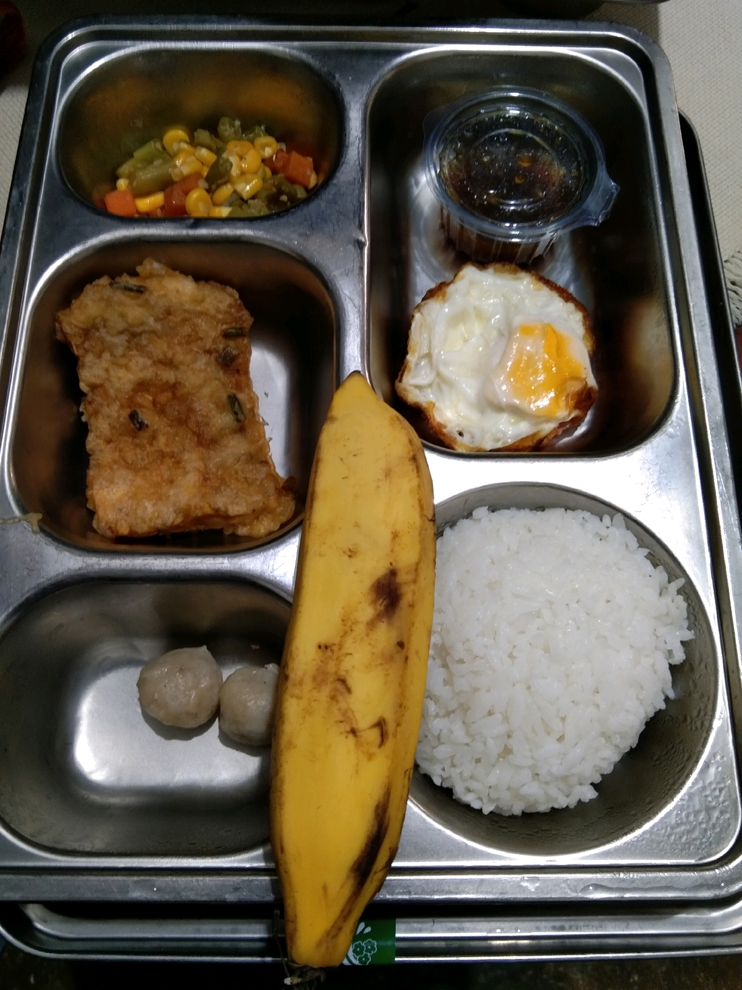 A segmented metal tray contains a meal. The top left compartment holds mixed vegetables including corn, green beans, and diced carrots. The top right compartment contains a small plastic container of dark liquid, likely a sauce or dressing, and a fried egg with a runny yolk. The middle left compartment has a piece of fried food, possibly a cutlet or a large fritter. The bottom left compartment contains two round meatballs or dumplings. A peeled banana lies horizontally in the center of the tray, overlapping the middle left and bottom compartments. The bottom right compartment is filled with white rice. The tray is placed on a surface that appears to be a table, with a woven texture visible. The image was taken from a high angle.
