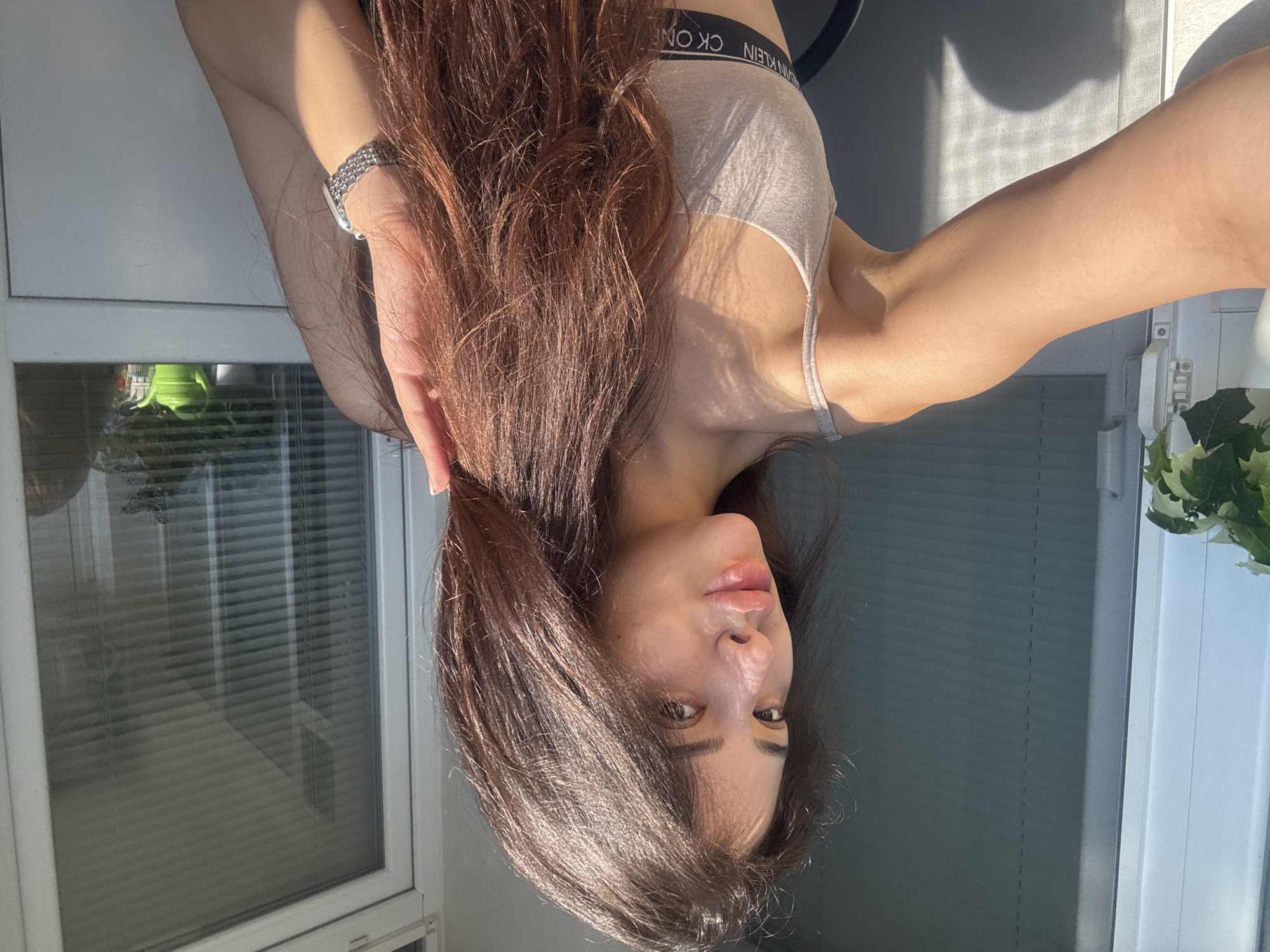 A young woman with long, dark brown hair and fair skin is captured in a selfie taken outdoors. She is wearing a light beige bralette with "CK ONE" visible on the elastic band. The woman's left arm is extended forward, holding what appears to be a phone, suggesting she is taking the photograph herself. Her right hand is positioned near her face, running through her hair. Sunlight casts shadows on her skin, highlighting her features, including full lips and luminous skin.
In the background, a window with horizontal blinds is visible, reflecting the ambient light. To the left of the window, there is a white structure with a green plant, and to the right, a small green watering can sits on the windowsill. The setting appears to be a balcony or patio, with a glimpse of a grey, textured wall behind the woman.
The image does not contain any specific identifiable landmarks that would pinpoint its location within the city of Plovdiv, Bulgaria.