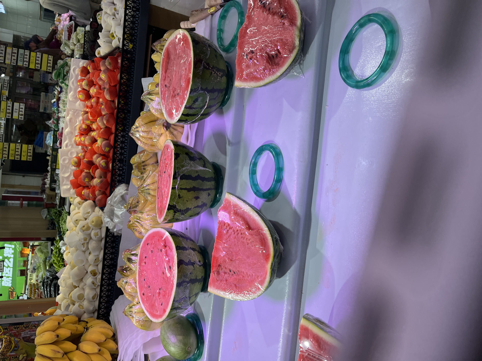 The image depicts a fruit and vegetable display at a market, possibly a grocery store, in Changchun City, China. A variety of produce is arranged in rows. Starting from the bottom and moving up, there are yellow bananas, white Asian pears, red peaches packed in red nets, and green leafy vegetables. On a white surface, there are cut watermelons in plastic wrap with green jade bangles next to them. Several price tags with Chinese characters are visible near the vegetables in the background.