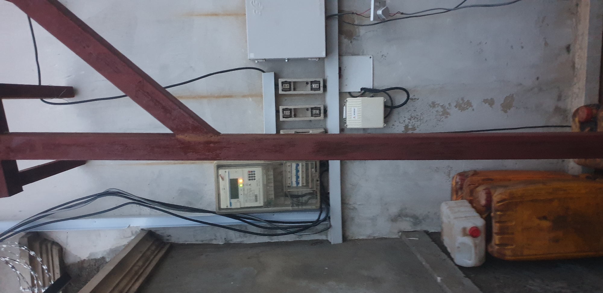 The image displays a utility installation on a weathered concrete wall in Narayi, Nigeria. In the foreground, a prominent reddish-brown metallic structural element, possibly part of a support frame or scaffolding, extends vertically and diagonally. To the left of this structure, an electricity meter is mounted on the wall. The meter is digital, featuring a display screen with text and numbers, a keypad, and two illuminated indicator lights: one green and one orange/red. Below the meter, circuit breakers are visible within a transparent enclosure. Several black electrical wires run vertically alongside the meter, partially contained within a light-colored conduit.
To the right of the metallic structure, further electrical components are installed. Two empty fuse holders or disconnect switches are mounted horizontally within a light-colored housing. Adjacent to these and to the right, a larger white rectangular electrical enclosure, marked with "EG," is affixed to the wall. Below the empty fuse holders, a smaller, cream-colored box with a triangular logo is connected by black cables. Towards the far right of the image, a white wall-mounted socket or junction box is visible with red and black wires connected.
At the bottom left of the scene, three plastic containers are stacked. Two larger, yellowish-orange jerrycans with red caps are at the base, and a smaller, white plastic container with a red cap rests on top. These containers appear used and somewhat dirty. In the upper left corner, strands of barbed or razor wire are visible, suggesting a security feature on a wall or fence above the depicted area. The concrete wall itself shows signs of wear, including dirt, discoloration, and some peeling paint or surface material, with rust-colored streaks visible in places.