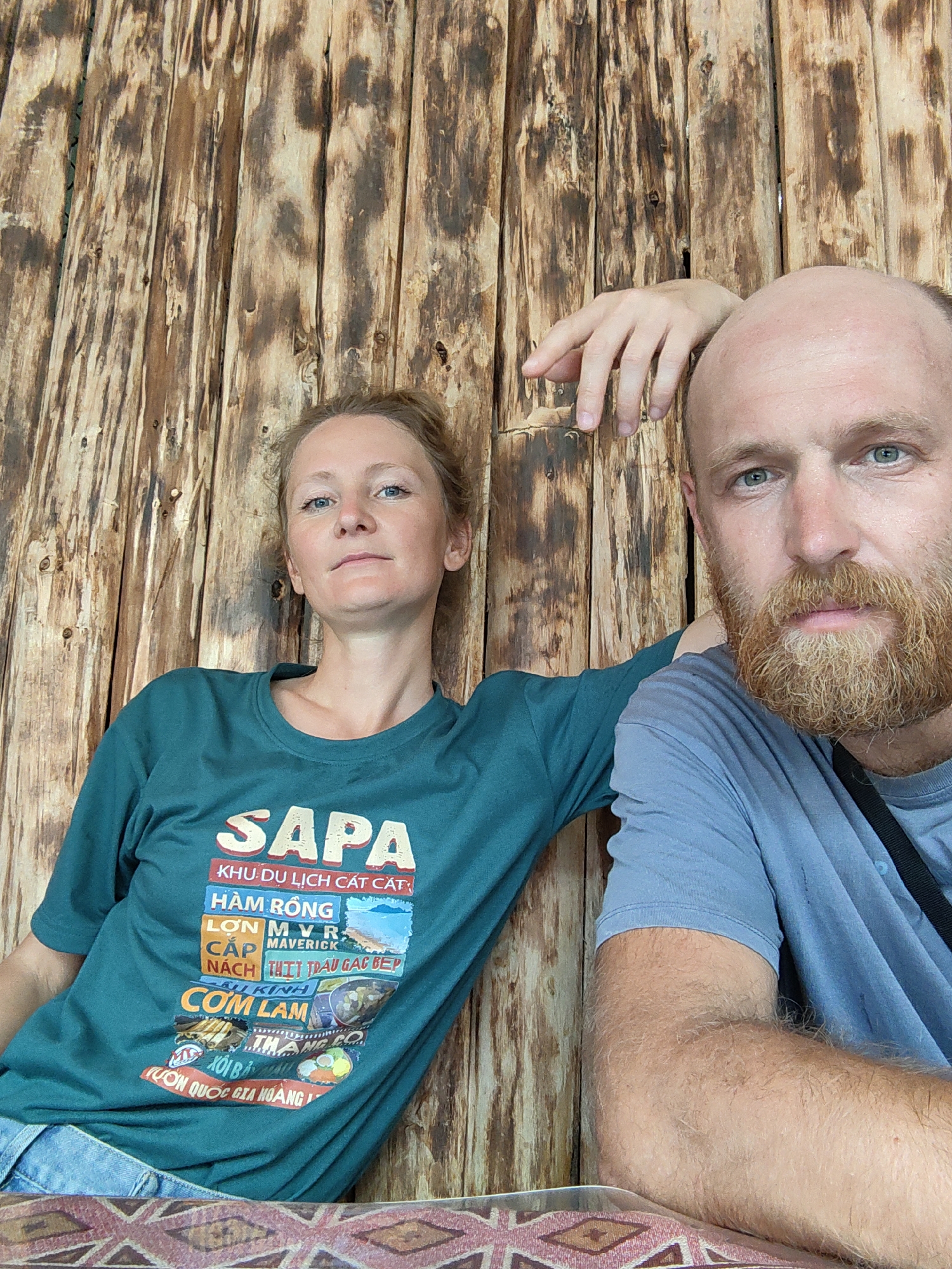 A man and woman are sitting close to each other in what appears to be a restaurant setting in Thành phố Hà Giang, Vietnam. They are posing for a photo, leaning against a wall constructed of rough, unfinished wooden planks. The woman is wearing a green t-shirt featuring a graphic design with the word "SAPA" prominently displayed at the top, followed by other text and images in Vietnamese relating to tourism in the region. The man is wearing a blue t-shirt. He has a shaved head and a red beard, and his arm is resting on the table in front of him. The table has a geometric patterned covering. The environment suggests they are in a rustic, possibly rural, location.