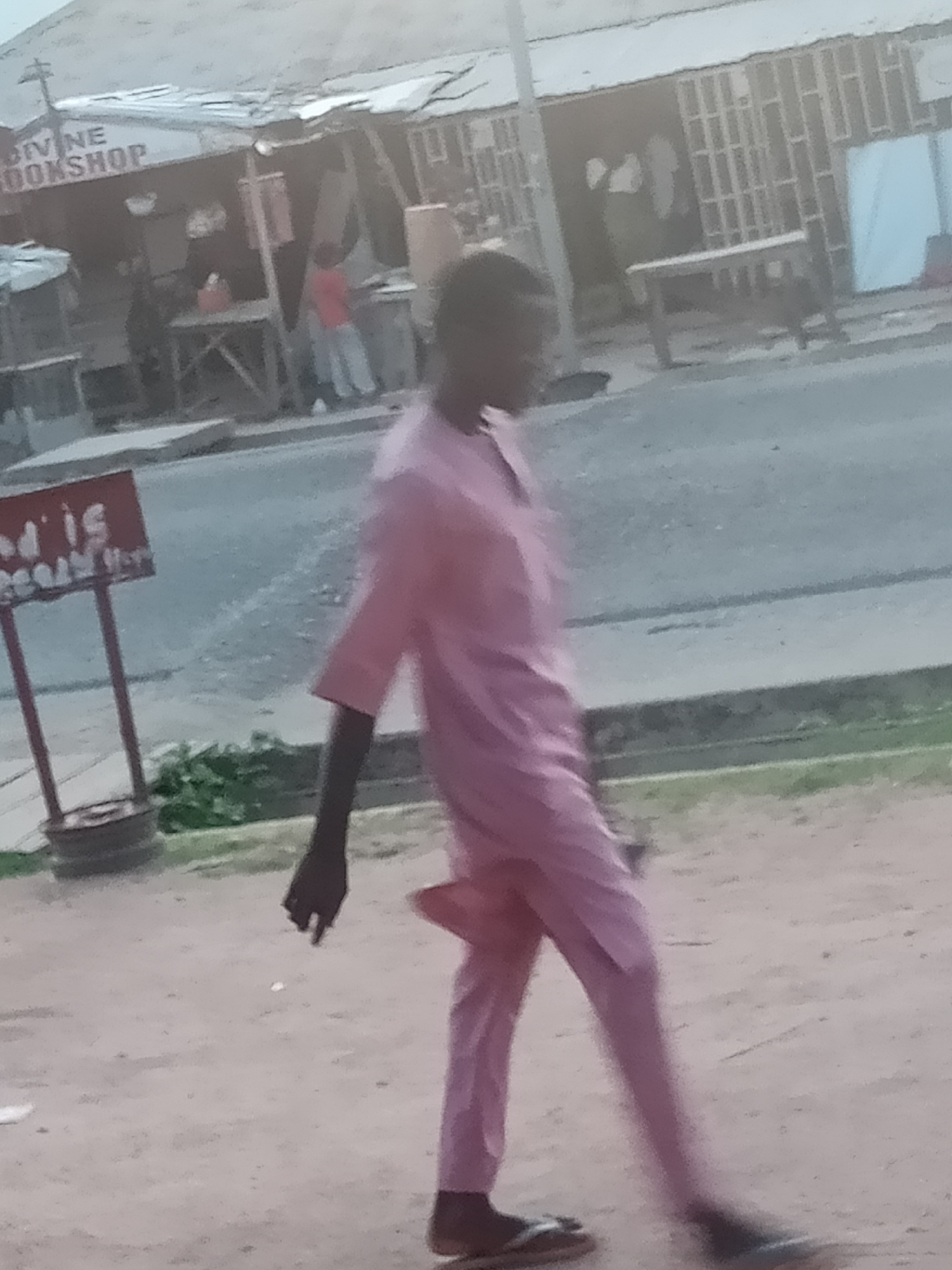A person wearing a pink robe and pants walks on a dirt road in Jalingo, Nigeria. The person is walking away from the camera and to the right. Behind the person is a small shop with a sign that reads "DIVINE BOOKSHOP". There are several tables and chairs outside the shop. The sky is overcast and the lighting is dim.