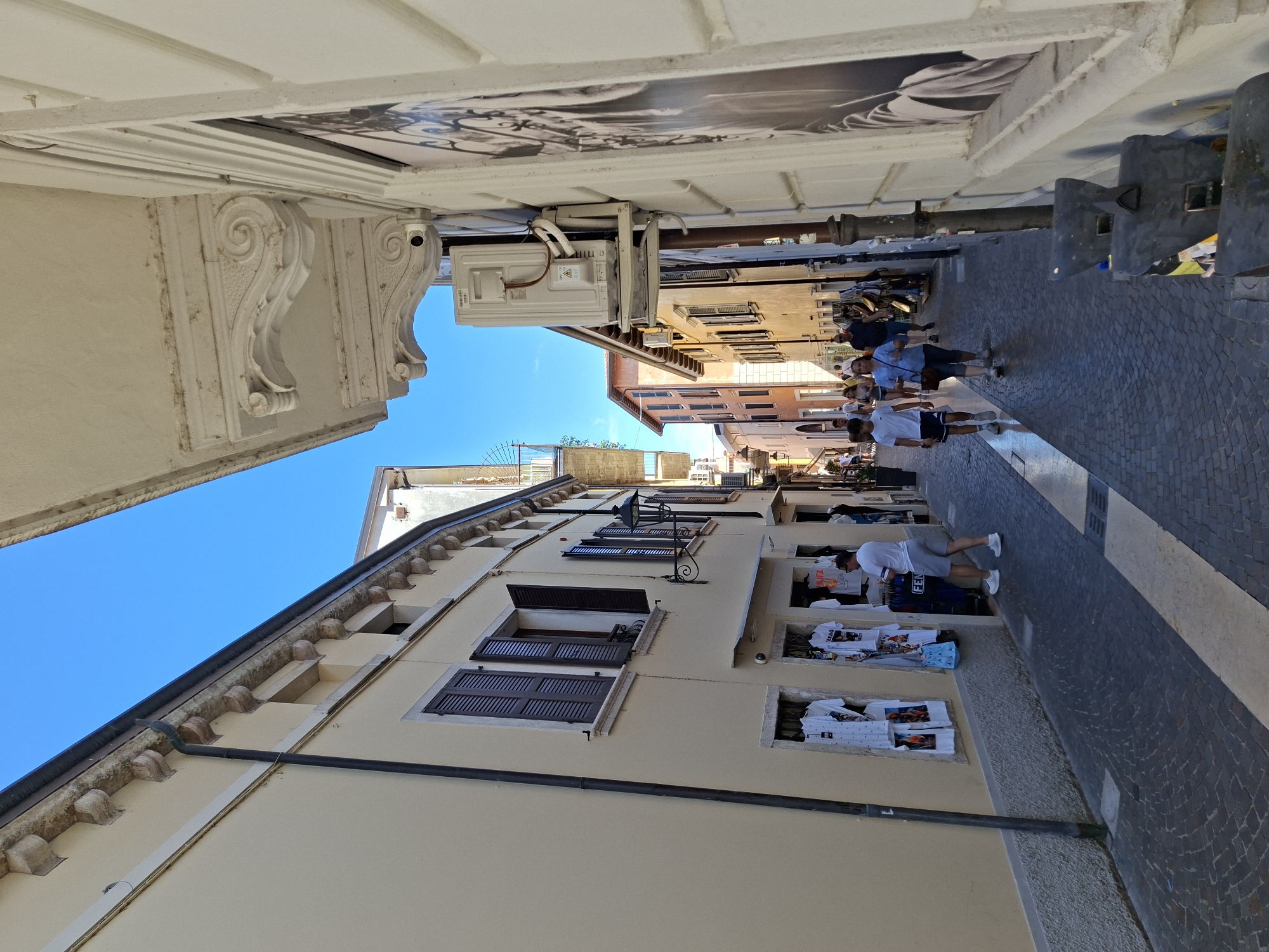 The image shows a narrow street in Peschiera del Garda, Italy, on a sunny day. The street is paved with cobblestones and lined with buildings on both sides. The building on the left is painted yellow and has several windows with dark shutters. There are racks of t-shirts displayed outside a shop on the ground floor. The building on the right is white and features decorative architectural elements, including ornate brackets supporting an overhang. An air conditioning unit is mounted on the exterior wall. Several people are walking along the street, some appearing to be tourists. The sky is clear and blue.
