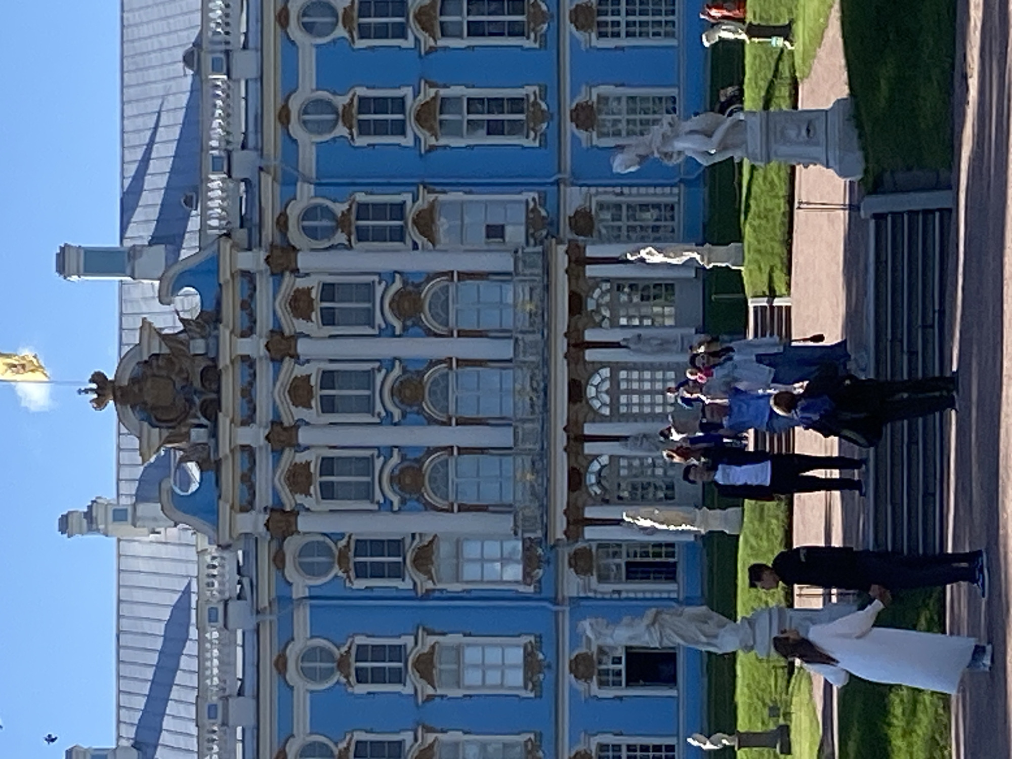 The image depicts the Catherine Palace in Pushkin, Russia, on a clear day with a bright blue sky. The palace is a large, ornate structure with a distinctive light blue facade, white trim, and gold accents. It features numerous windows, columns, and decorative elements. In the foreground, several people are visible. A couple is holding hands on a path in front of the palace steps. A man in a black jacket and white t-shirt stands on the steps, with his arm raised. Other individuals are scattered throughout the scene, some walking and some standing. Several statues are placed on pedestals on the lawn surrounding the palace. The environment is well-maintained, with manicured green lawns and paved pathways. A yellow flag flies from a flagpole atop the palace.