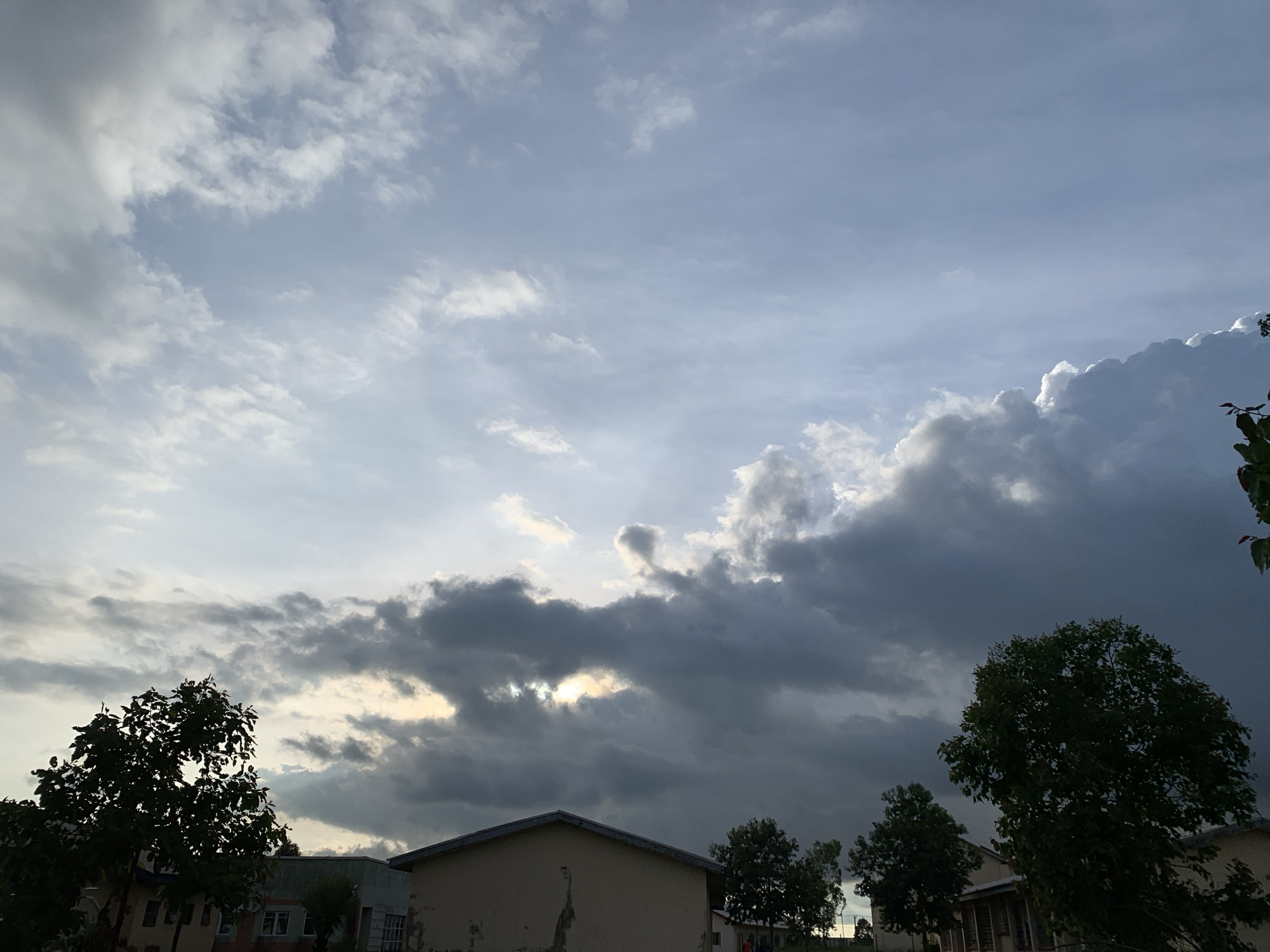 The image shows a view of the sky and rooftops in Kuru Karama, Nigeria. The sky is filled with a dramatic array of clouds, with dark, heavy storm clouds dominating the lower right portion of the frame and lighter, more scattered clouds in the upper left. Patches of sunlight break through the clouds in the center, creating a dramatic contrast. Silhouetted against the sky are the tops of several trees, their leaves appearing dark green. In the foreground, the roofs of buildings are visible, appearing to be residential structures. The overall impression is that of an impending storm or the aftermath of one. No people or specific actions are depicted in the scene.