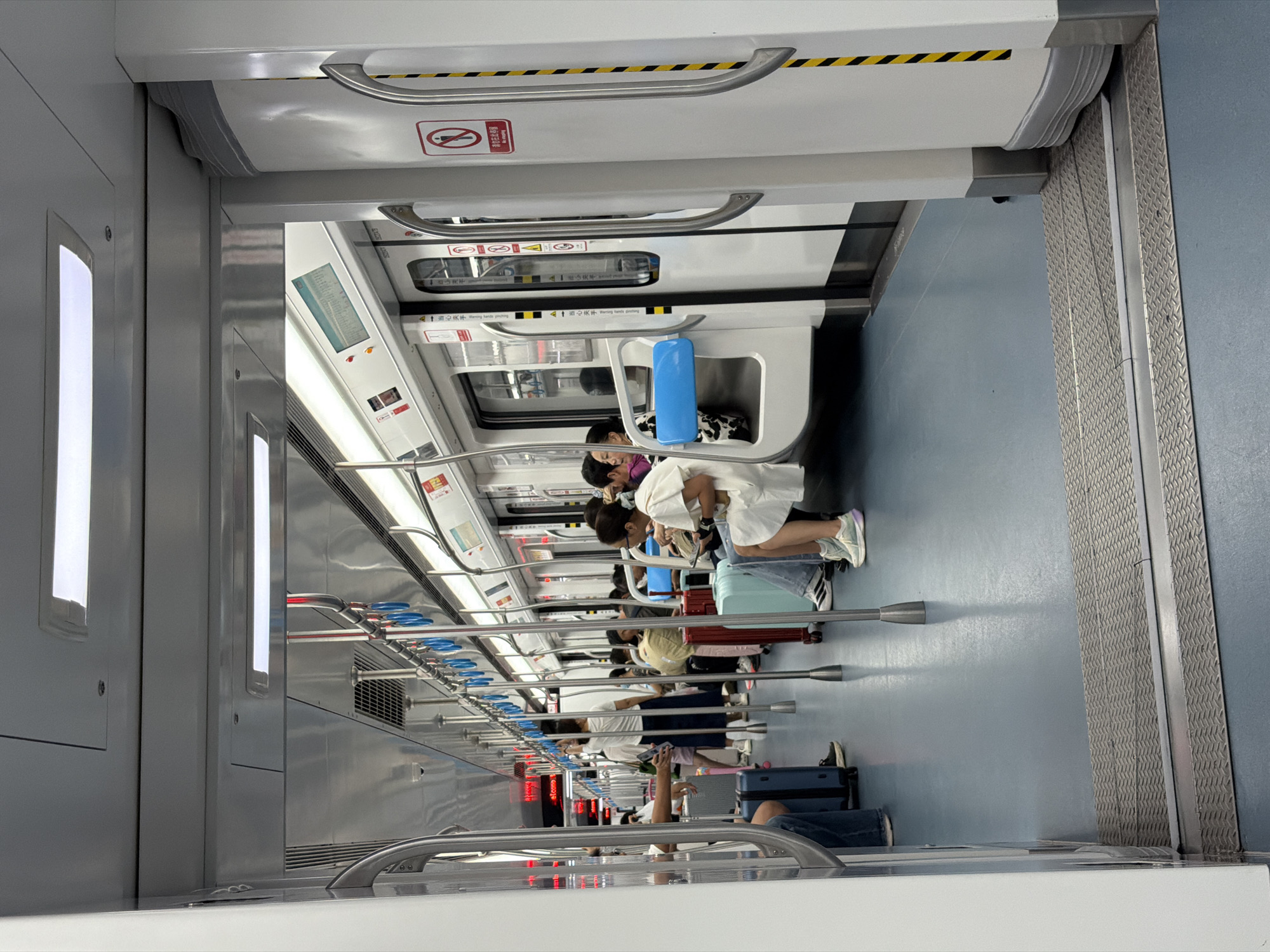 An interior view of a subway car shows passengers seated and standing. The car has light blue flooring and white walls with silver poles for passengers to hold onto. Several passengers are seated on the bench-style seats, with some having luggage next to them. A woman in a white dress is seated on the right side of the car, and other passengers are visible in the background, some standing and holding onto the poles. The lighting appears to be from overhead fluorescent fixtures. The scene suggests a typical commute or travel situation within a public transportation system.