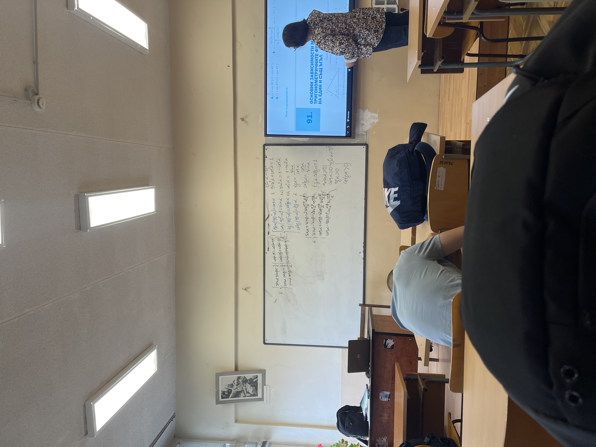 This image captures a classroom scene in Sofia, Bulgaria, from a student's perspective. In the foreground, the back of a student's head and shoulder, wearing a black jacket, is visible. Seated in front of this student, another individual wears a light blue t-shirt, also seen from the back. A blue backpack with a prominent "NIKE" logo rests on a chair next to a wooden desk, which has "957496" printed on it.
At the front of the classroom, a woman, possibly a teacher, stands near a large digital display screen and a whiteboard. She is wearing a floral-patterned top and appears to be interacting with the screen. The screen displays Bulgarian text and mathematical content related to trigonometry, including headings like "ОСНОВНИ ЗАВИСИМОСТИ М" (MAIN DEPENDENCIES) and "ТРИГОНОМЕТРИЧНИТЕ ФУТ НА ЕДИН И СЪЩ ЪГЪЛ" (TRIGONOMETRIC FUNCTIONS OF ONE AND THE SAME ANGLE), along with specific problems and "91." The whiteboard is covered with numerous handwritten trigonometric identities and formulas in black marker, such as "sin(90°-α) = cos α" and "sin²α + cos²α = 1", along with conditions like "sin α > 0".
The classroom features light-colored, textured walls, with rectangular fluorescent light fixtures illuminating the room from the ceiling. A white smoke detector is also visible on the ceiling. A framed picture hangs on the wall between the whiteboard and a window. The floor has visible wooden parquet. The scene depicts an active lesson during what appears to be daytime, given the bright indoor lighting.