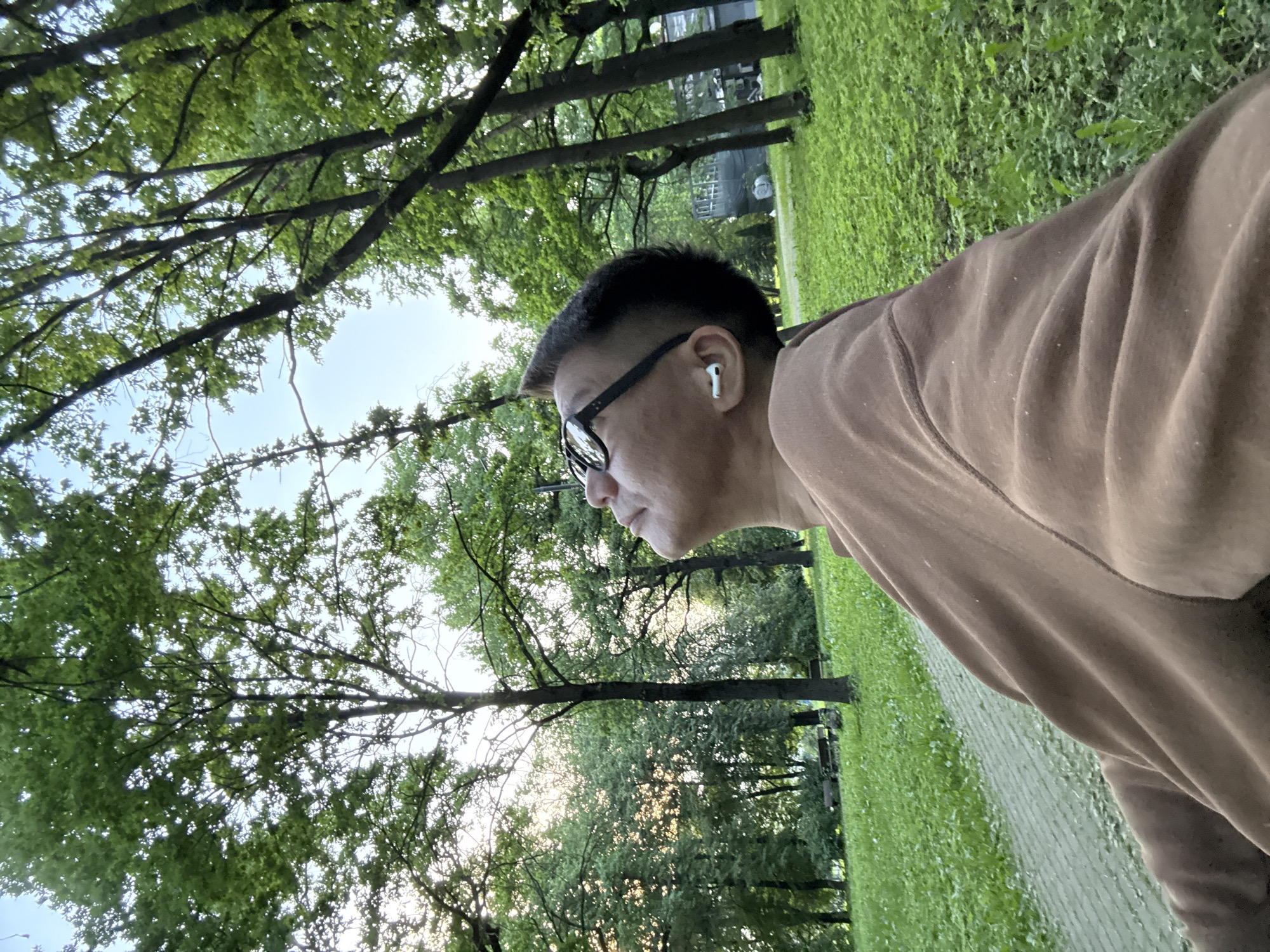 A man wearing glasses and white earbuds is looking up at the sky in a park. He is wearing a brown sweatshirt. The park has green grass, trees, and a paved path. There are benches in the background. The sun is setting, casting a warm glow on the trees. The location is the city of Khimki, Russia.