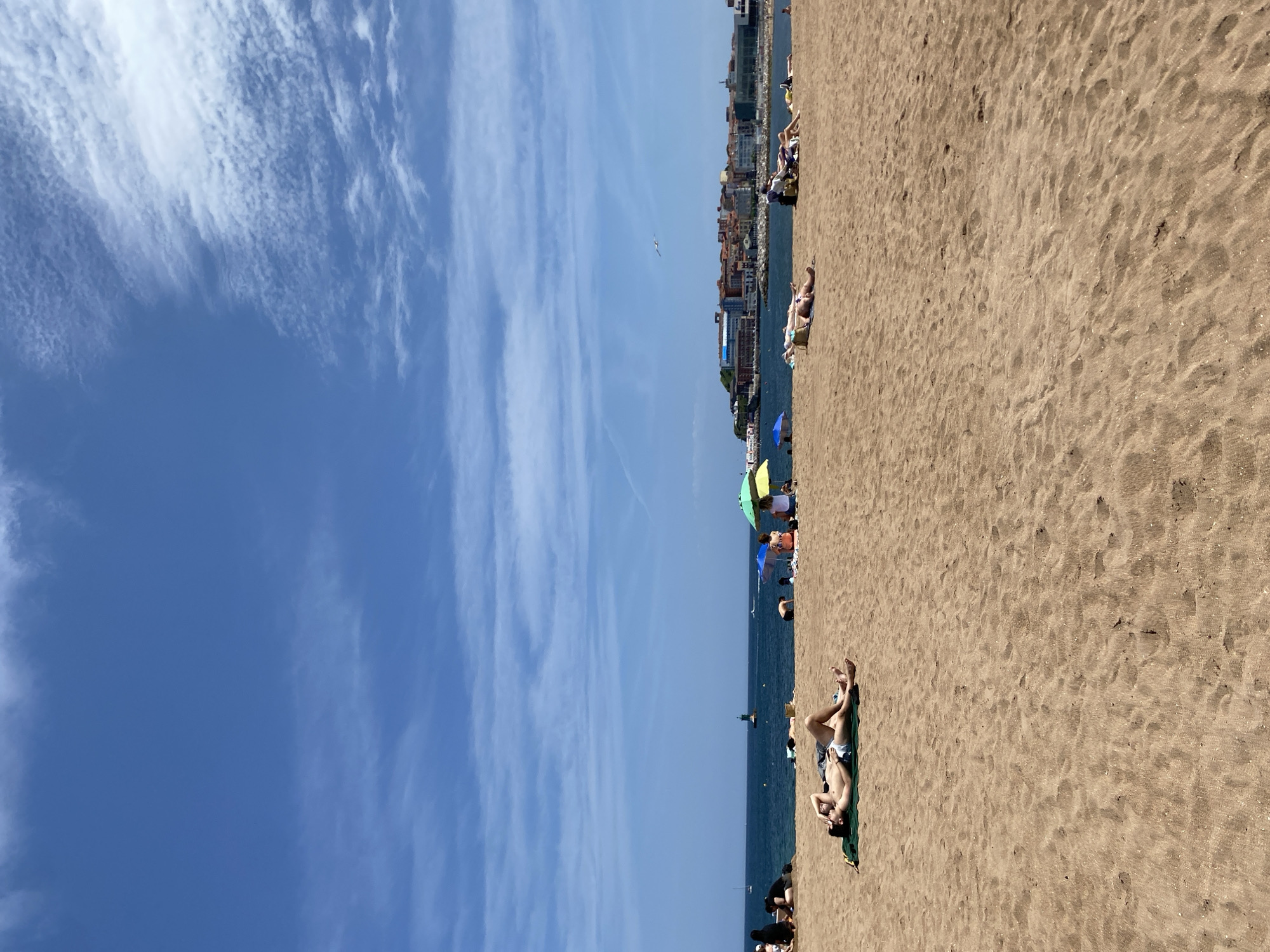 This picture was taken at a beach in Gijón/Xixón, Spain. The picture shows a beach with several people on it. Some are lying down sunbathing, while others are sitting under umbrellas. There is a clear blue sky with wispy white clouds and a calm sea. Buildings are visible in the distance.