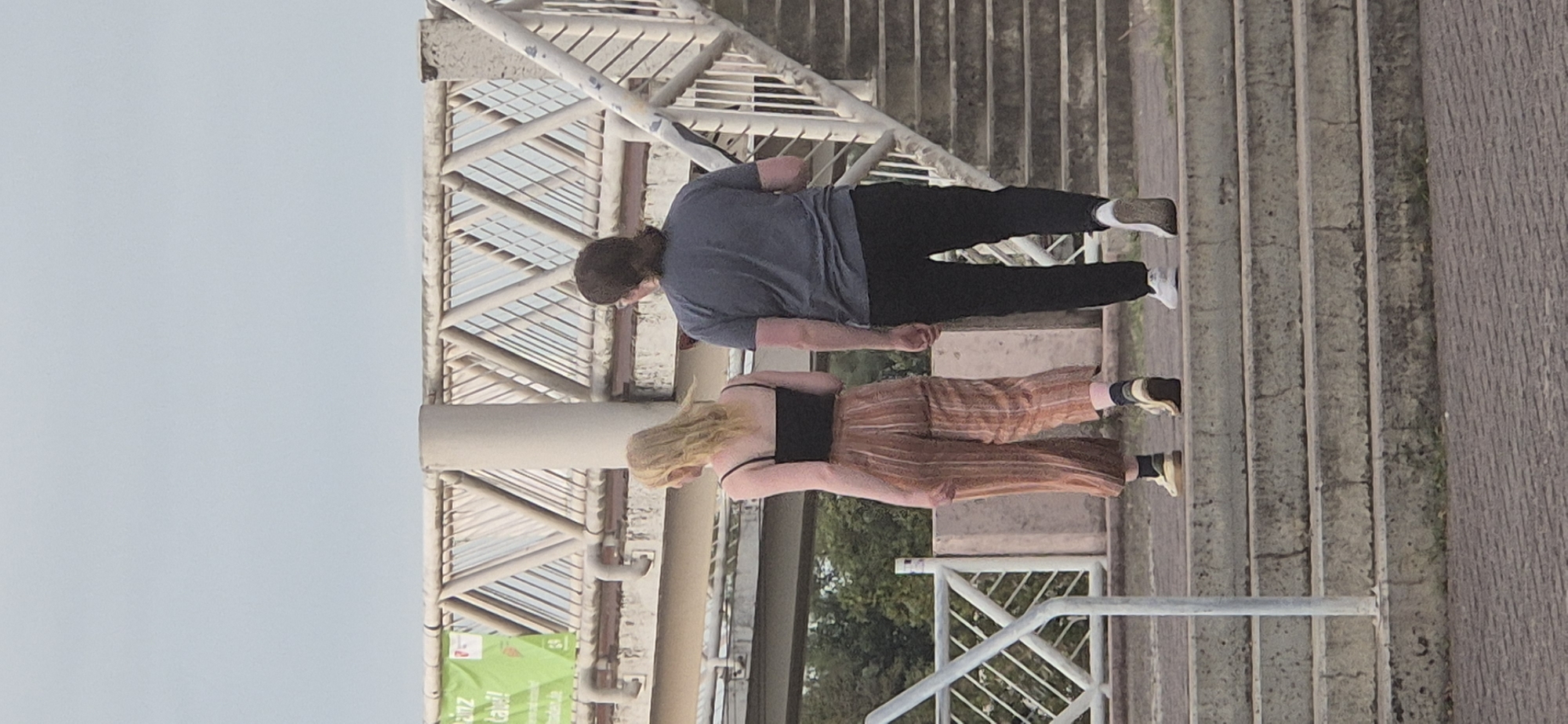 Two individuals, viewed from behind, are walking up a concrete staircase. The person on the left has blonde hair and is wearing a black tank top and striped orange pants. The person on the right has dark, shoulder-length hair and is wearing a dark grey t-shirt and black pants. They are on a pedestrian bridge. In the background, a green banner with German text, possibly advertising a local event or establishment, is visible. The overall scene suggests an outdoor public space, likely within the city of Mainz, Germany, given the presence of a bridge and urban infrastructure.