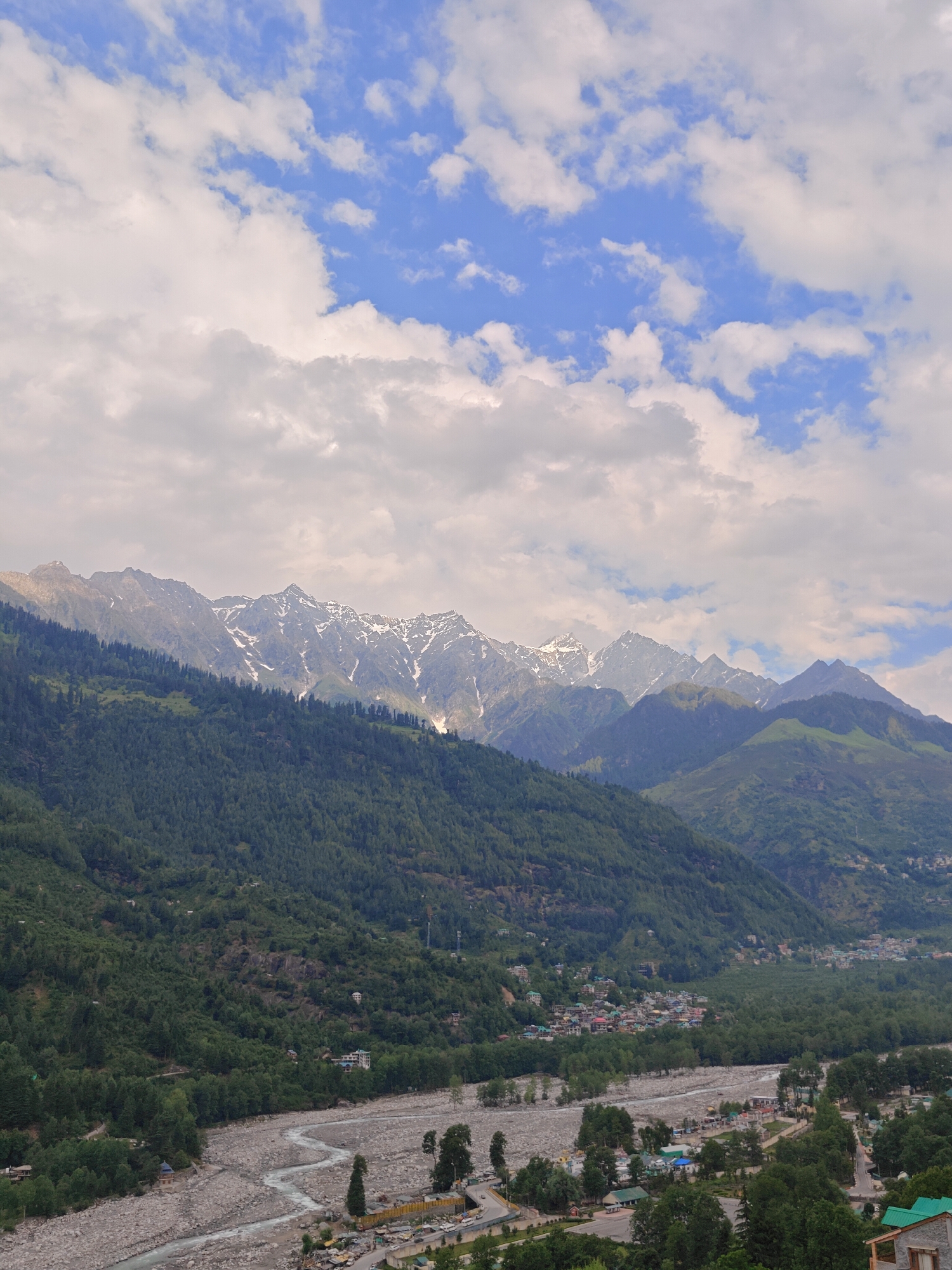 The image depicts a wide, scenic view of mountains near Vashisht, India, under a partly cloudy sky. In the foreground, a wide, rocky riverbed with a meandering, shallow river dominates the lower portion of the frame. A small settlement with buildings and roads is visible along the riverbank, nestled amongst lush green trees. As the view moves upward, the landscape transitions into steep, densely forested mountainsides. The upper slopes of the mountains reveal rugged, rocky peaks, some of which are capped with snow, indicating a high altitude. The sky is a mix of blue patches and white, fluffy clouds. The scene captures a natural landscape with a human settlement integrated into it, suggesting a rural or semi-urban mountain environment.