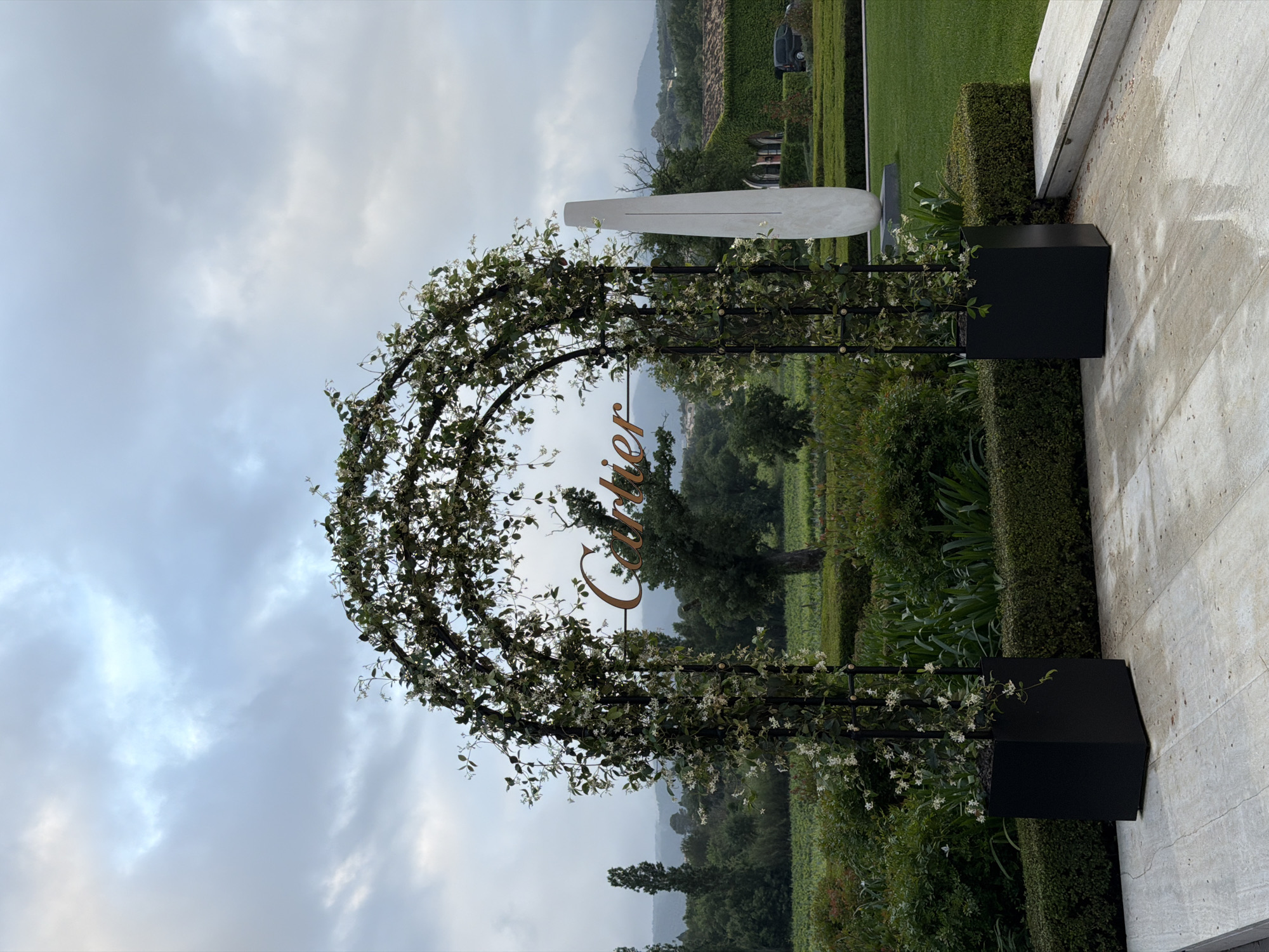 The image shows an outdoor setting, likely a garden or vineyard, near Cogolin, France. An archway, decorated with climbing plants bearing small white flowers, frames a view of the landscape. The word "Cartier" is mounted in a script font on the archway. The arch is supported by black rectangular planters on each side. Beyond the arch, the landscape features greenery, including trees, hedges, and cultivated fields. In the background are rolling hills obscured by an overcast sky. To the right of the archway is a modern white sculpture and a wall with a light stone finish. The ground is damp, likely from recent rain.