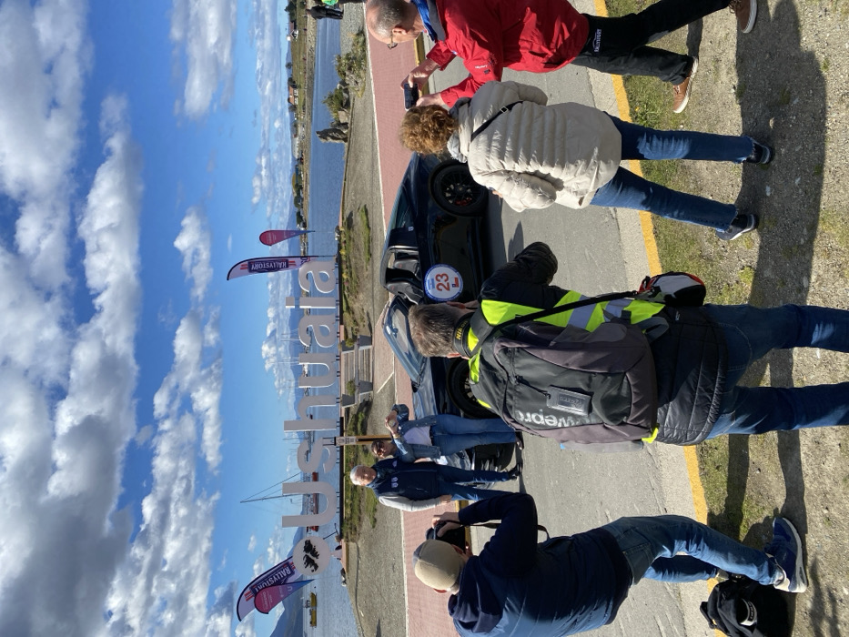 The scene depicts an outdoor gathering in Ushuaia, Argentina, on a sunny day with a partially cloudy blue sky. In the background, a large white "Ushuaia" sign, incorporating a stylized heart with the flag of the Falkland Islands/Malvinas, is prominently displayed near a body of water. Several sailboats are visible in the water, and distant landmasses line the horizon. Two purple and white "RALLYSTORY" feather flags are erected on the waterfront, indicating an event.
In the midground, a black two-door performance car, identified by a white circular "23 L" sticker on its side, is parked on a reddish-brown paved surface next to a curb. A man and a woman stand in front of the car, posing for photographs. The man wears a navy vest over a white shirt and jeans, while the woman wears a denim jacket and jeans, forming a heart shape with her hands.
In the foreground, four individuals are engaged in photography. To the left, a man in a dark blue jacket and beanie is crouched, holding a black DSLR camera to his eye. To his right, another man wearing a yellow reflective vest and a "LOWEPRO" backpack is also holding a black "Nikon" DSLR camera, aiming it at the posing couple and car. To the far right, a woman in a light beige puffer jacket and a man in a red jacket with "RALLYSTORY" branding on his pants are both holding smartphones horizontally, taking pictures of the scene. A black backpack rests on the ground near the left photographer. The ground is a mix of paved surface and light-colored gravel/dirt, with shadows cast by the people.