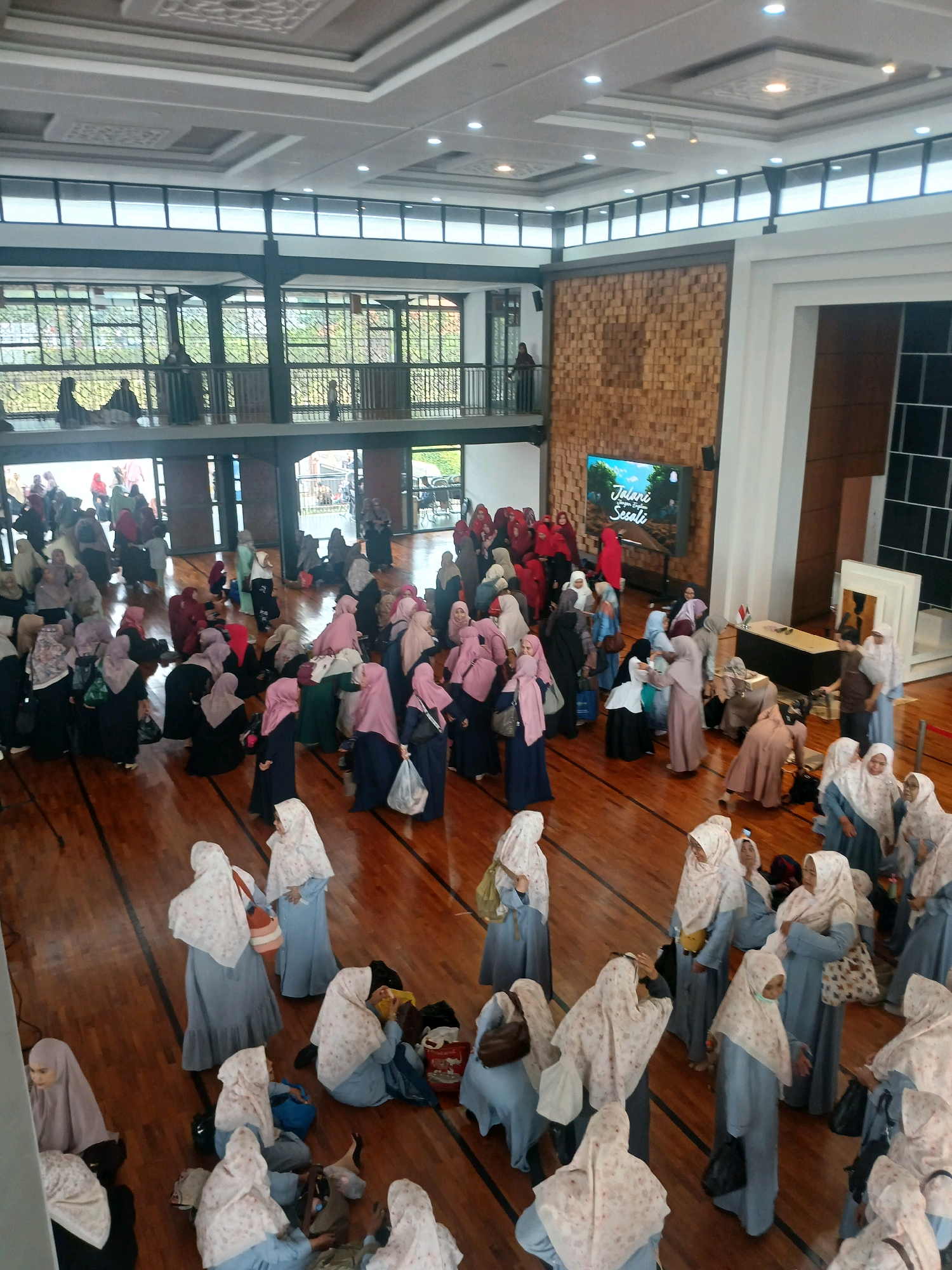 The image depicts a large indoor gathering of people, predominantly women, in what appears to be a community hall or event space with a wooden floor. The individuals are dressed in a variety of traditional Islamic attire, including hijabs and long dresses in colors such as blue, pink, white, and black. Many are standing and conversing, while some are seated on the floor, with bags and belongings around them.
In the background, there is a large screen displaying an image with text that reads "Jalani Sesuatu". Adjacent to the screen is a wall with a distinctive textured wooden paneling. A balcony area with railings and glass enclosures is visible in the upper portion of the image, with more people gathered there. Natural light enters the space from windows and overhead lighting fixtures illuminate the interior. The overall atmosphere seems to be one of a social or religious event.