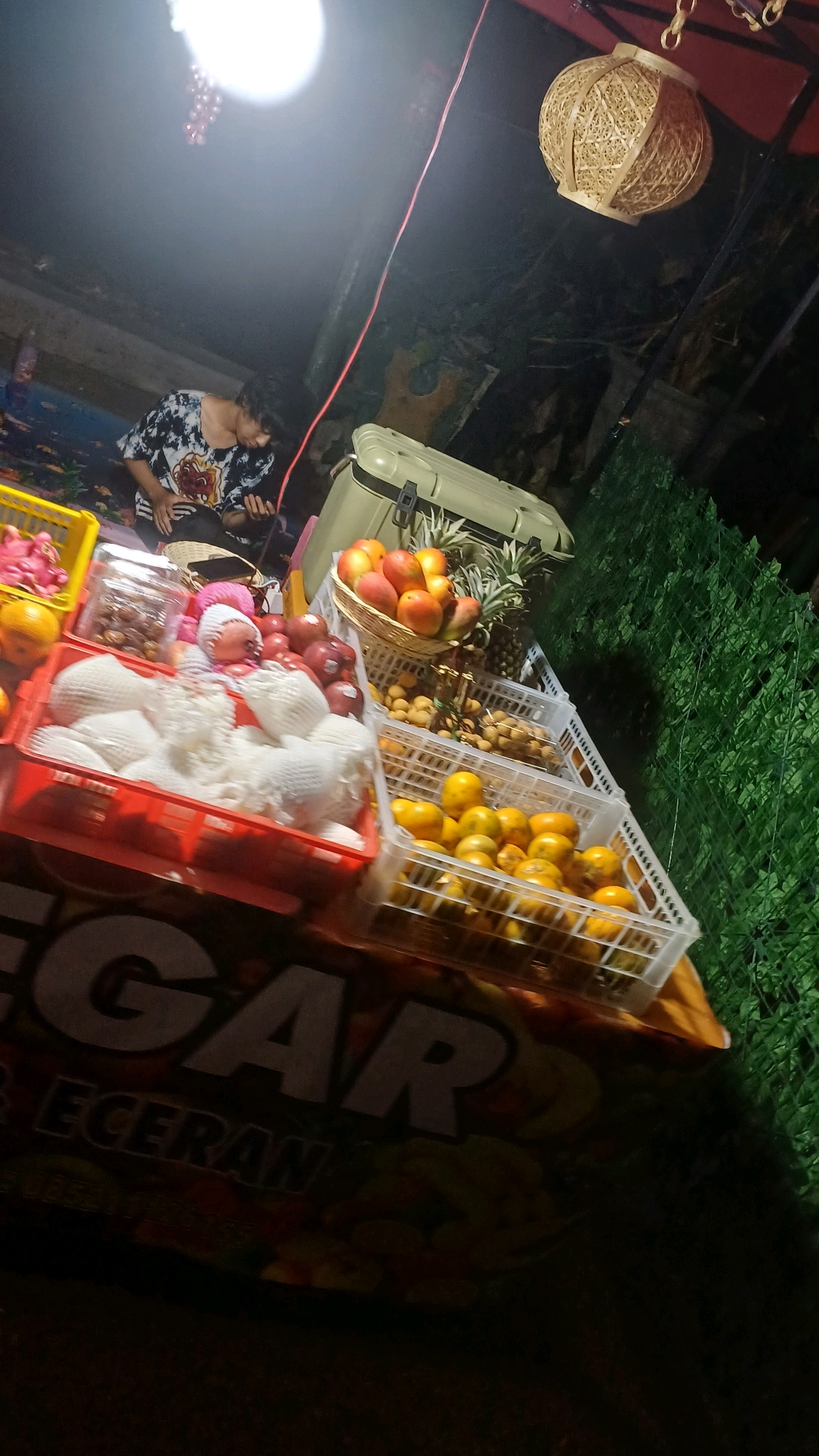 A person is sitting on the ground in front of a fruit stand at night in Kowangan, Indonesia. The stand is decorated with a spherical woven lantern hanging from above. The person is wearing a black and white patterned shirt and is looking down at their phone.
The fruit stand is laden with various fruits, including mangoes in a basket, pineapples, red apples, and what appear to be pears or similar white-fleshed fruit individually wrapped in white foam netting. Below the fruits, a large banner with text in what appears to be Indonesian is visible, with the word "SEGAR" prominent.
The background shows a wall covered in green artificial foliage. A light source illuminates the scene from above and to the left, casting shadows and creating a dramatic nighttime atmosphere. A cooler is also visible behind the fruit display. The overall scene depicts a typical nighttime market setting in Indonesia.