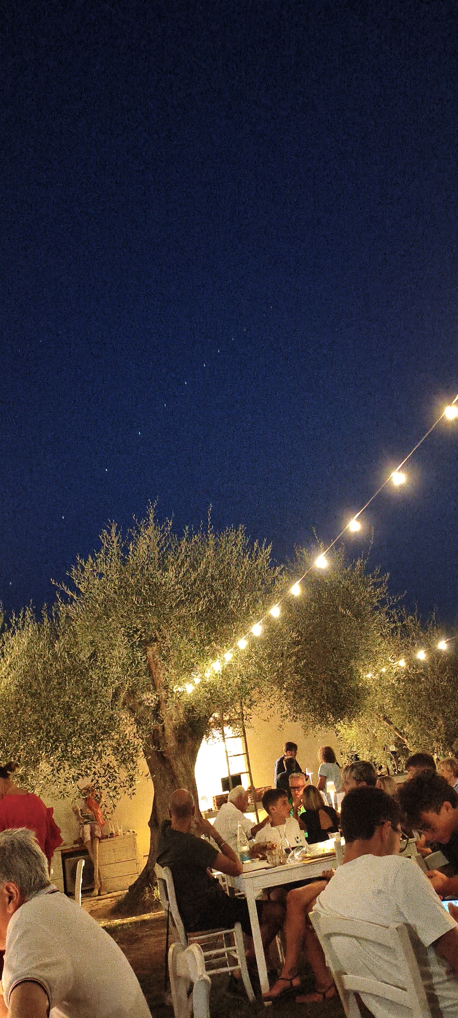 This is a nighttime photograph of an outdoor gathering in Buriano, Italy. The scene depicts a group of people seated at tables, enjoying a meal or drinks under a string of illuminated lights. The background features olive trees and a building, suggesting a rural or semi-rural setting. The sky is dark blue, with a few faint stars visible. The overall atmosphere appears relaxed and convivial.