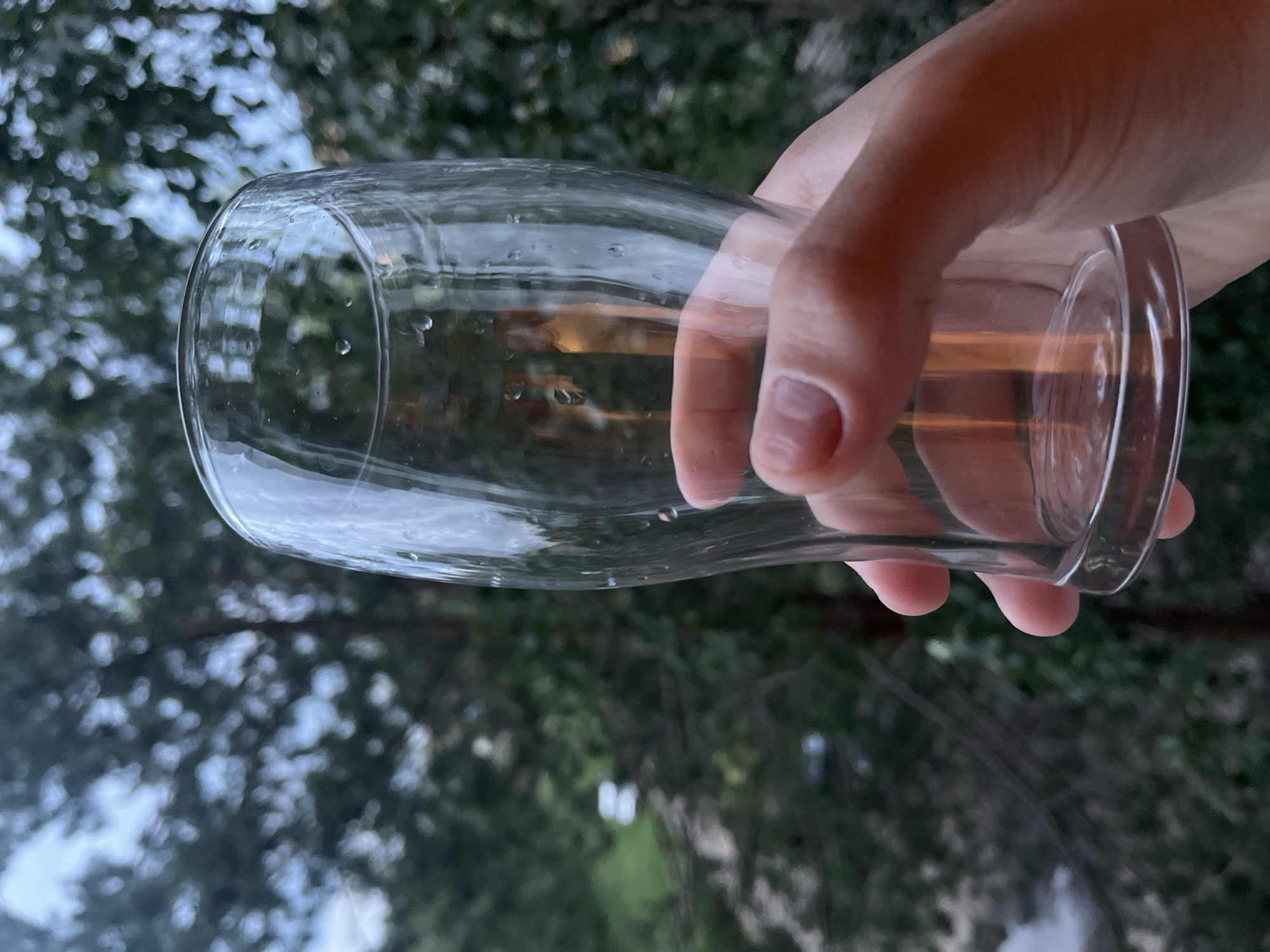 A person's hand holds a clear glass with water droplets on its exterior. The glass is positioned in the foreground, with a blurred background of trees and foliage. The lighting suggests it is daytime, possibly evening, with soft, diffused light. The location is unknown, and the scene appears to be outdoors, perhaps on a balcony or near a window with a view of nature.