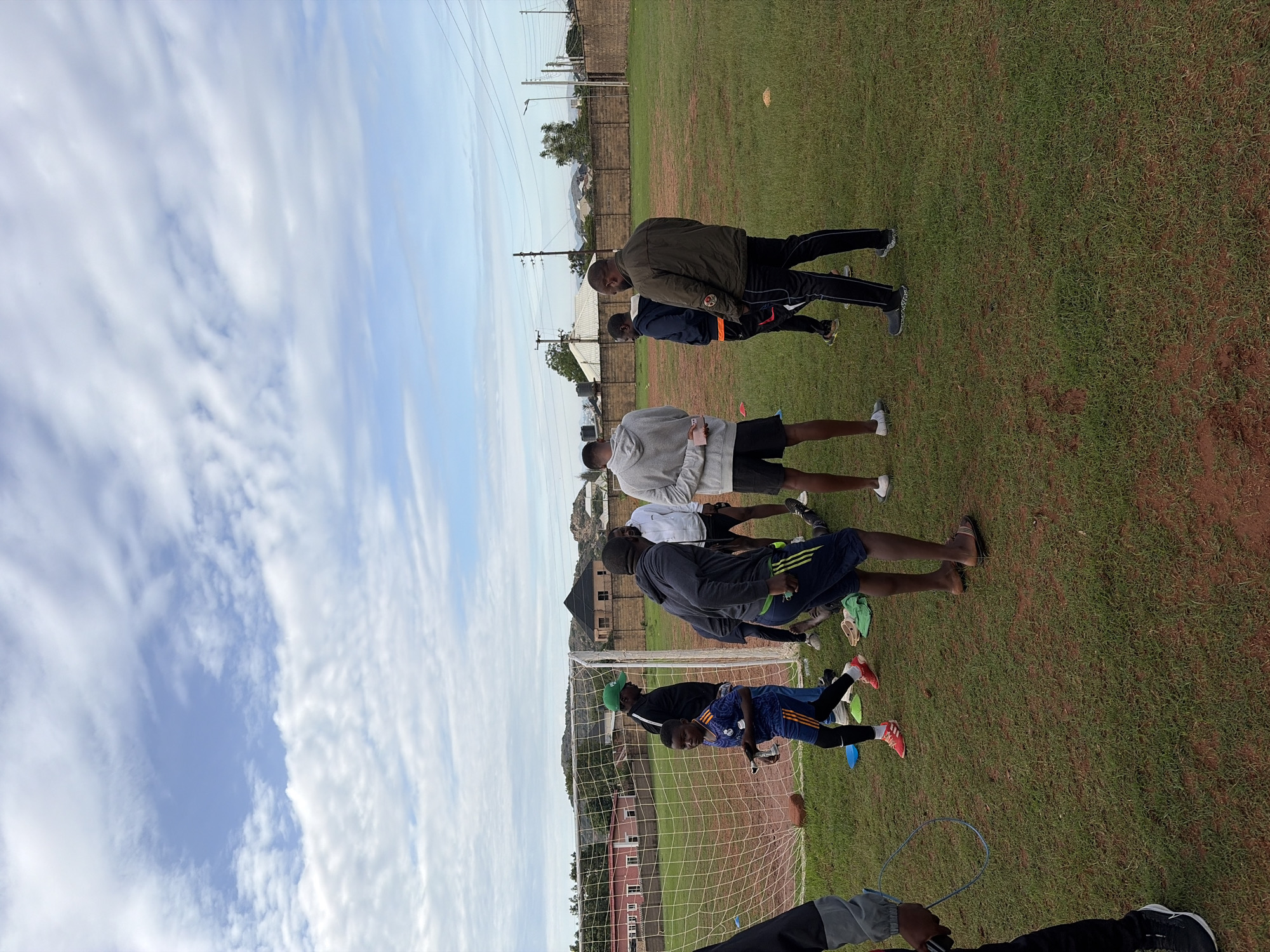 The image depicts an outdoor sports field in Jos, Nigeria, characterized by its mix of green grass and reddish-brown earth. At least nine individuals are present, consisting of adult males and one young boy, suggesting a training session or informal gathering.
In the left foreground, a person, largely out of frame, wears a dark jacket, black pants, and sneakers, holding a blue skipping rope. To their right, a young boy in a blue and orange Real Madrid football kit and red-and-white sneakers stands near a white goalpost with a net, holding a small silver object. Behind him, a man wearing a green cap, black jacket, and blue jeans stands by the goalpost, observing the group.
In the central area, a cluster of men is visible. One man in a grey hooded sweatshirt and black shorts stands with his back to the viewer, positioned next to another man in a white hooded sweatshirt and black shorts, also facing away. To their right, an adult male, potentially a coach, wears a dark green/khaki jacket and black track pants with light stripes, facing left as if addressing or observing the others. Another man in a blue jacket and black shorts with orange stripes stands facing the center-right, engaging with the group. Partially obscured on the far right, another person's legs are visible in white shoes. Two pairs of slippers are placed on the grass in the mid-ground. Small orange cones are scattered on the field, indicating potential training markers.
The background features residential buildings with red roofs and high perimeter walls, interspersed with scattered trees. Distinctive hills or mountains, characteristic of the Jos Plateau, rise in the distant background. Utility poles and overhead wires are visible on the right side of the frame. The sky is partly cloudy with patches of clear blue visible.