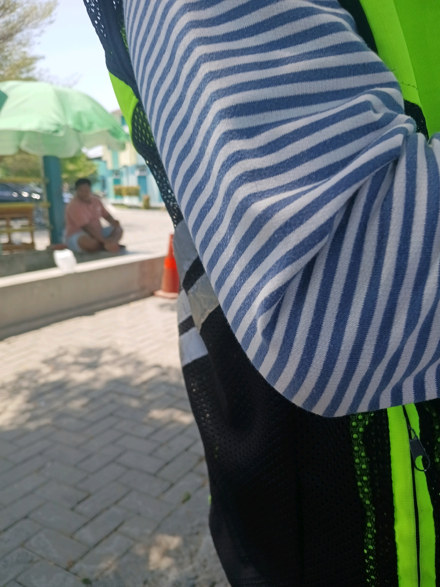 The image shows a close-up of a person wearing a striped blue and white long-sleeved shirt over a black and neon green vest. The vest has a mesh texture and a zipper. The background is blurred and appears to be an outdoor setting. A person is sitting on a ledge in the background, wearing a pink shirt and light blue shorts, and an open green umbrella is behind them. The ground is paved with grey interlocking bricks. The overall impression is a casual outdoor scene, possibly in a park or public area within the city of Unknown, Unknown.