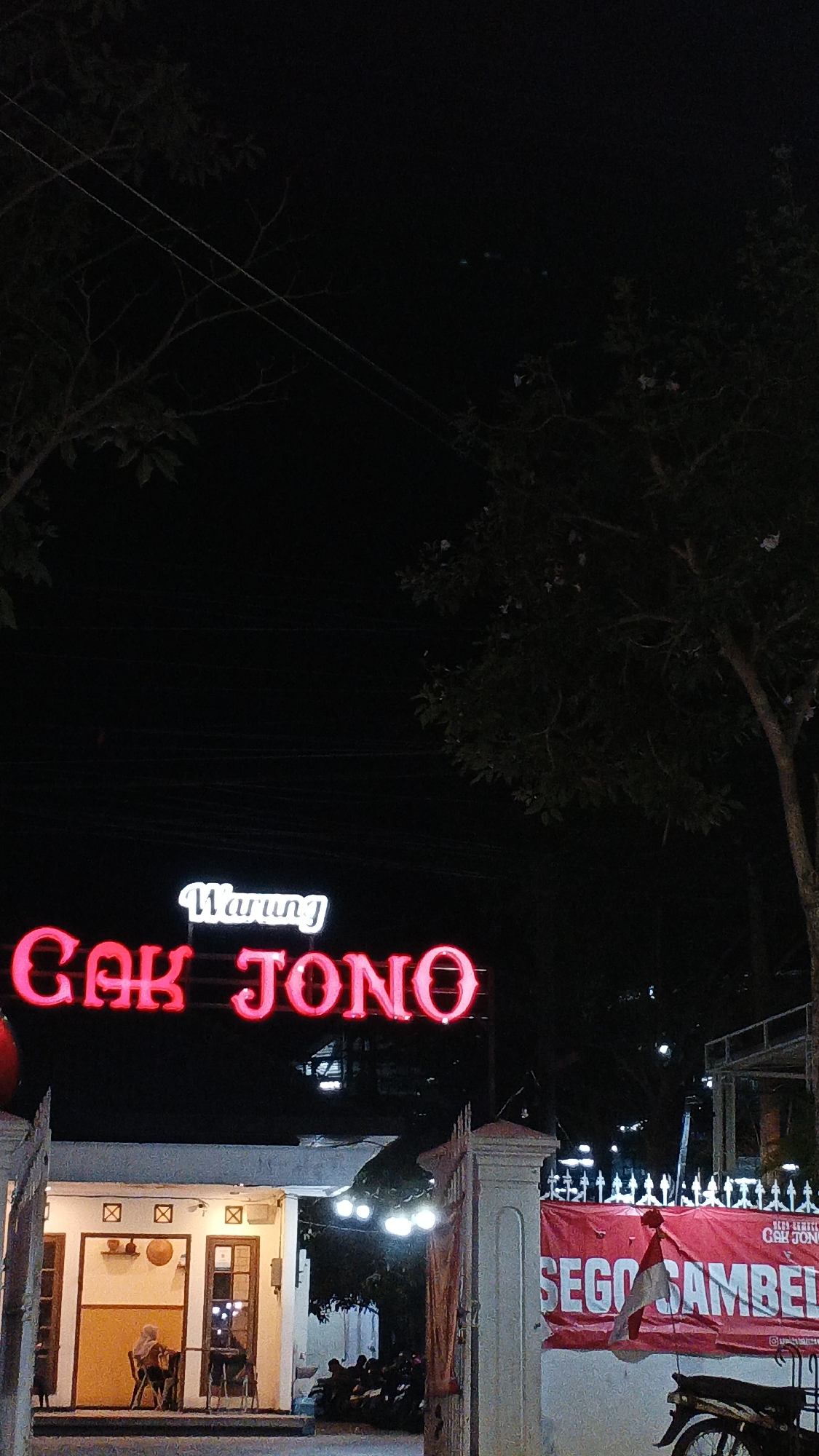 A neon sign that reads "Warung Cak Jono" is illuminated in red and white against a dark night sky. Below the sign, a building with an open-air entrance is visible, with a few people seated inside and outside. A red banner with white text that appears to read "Sego Sambel" is hanging on a pole to the right of the building, partially obscuring a motorcycle. The scene suggests a casual dining establishment, likely in a moderately populated area, given the presence of streetlights and a few parked motorcycles. The overall ambiance is that of a bustling evening eatery.